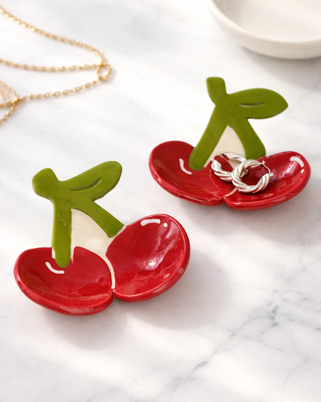 Ceramic cherry ring dish holding silver hoop earrings on marble surface with gold necklace, fruit shaped jewelry tray for vanity organization