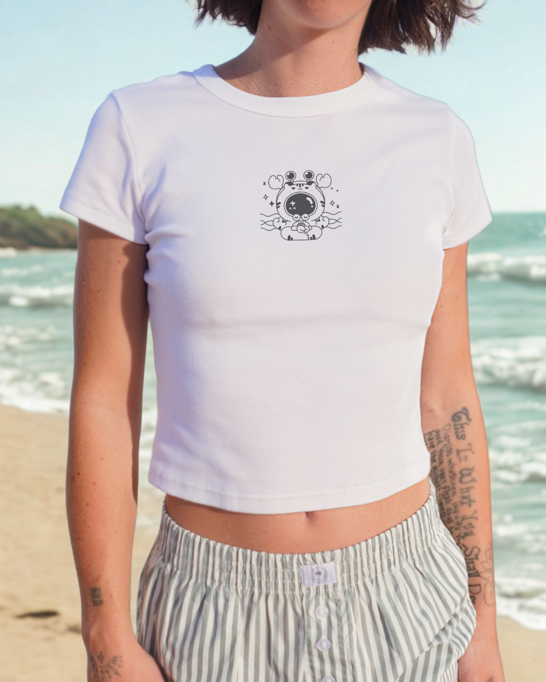 Cropped white baby tee with meditating crab illustration styled in a coastal outdoor setting