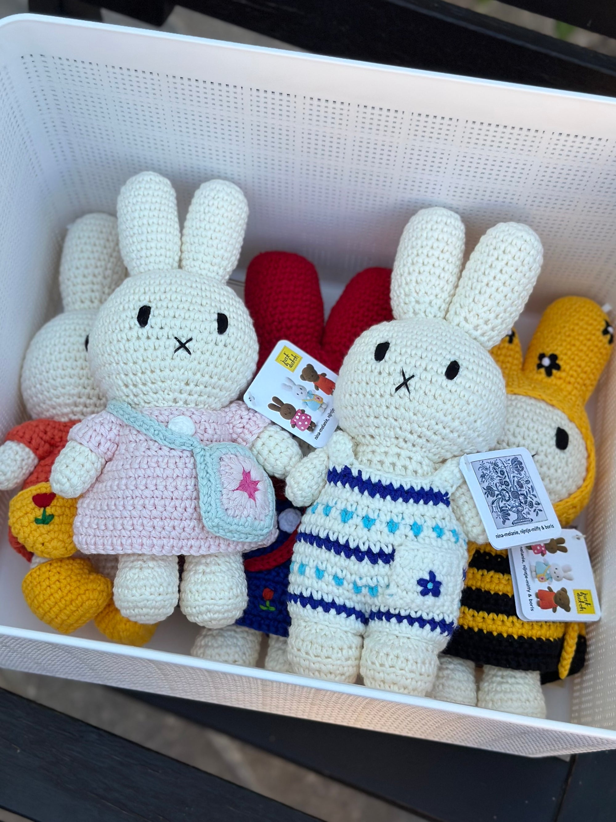 Handmade Miffy crochet plush in Delft blue floral jumpsuit sitting in a basket with other crochet Miffy dolls in colorful outfits.