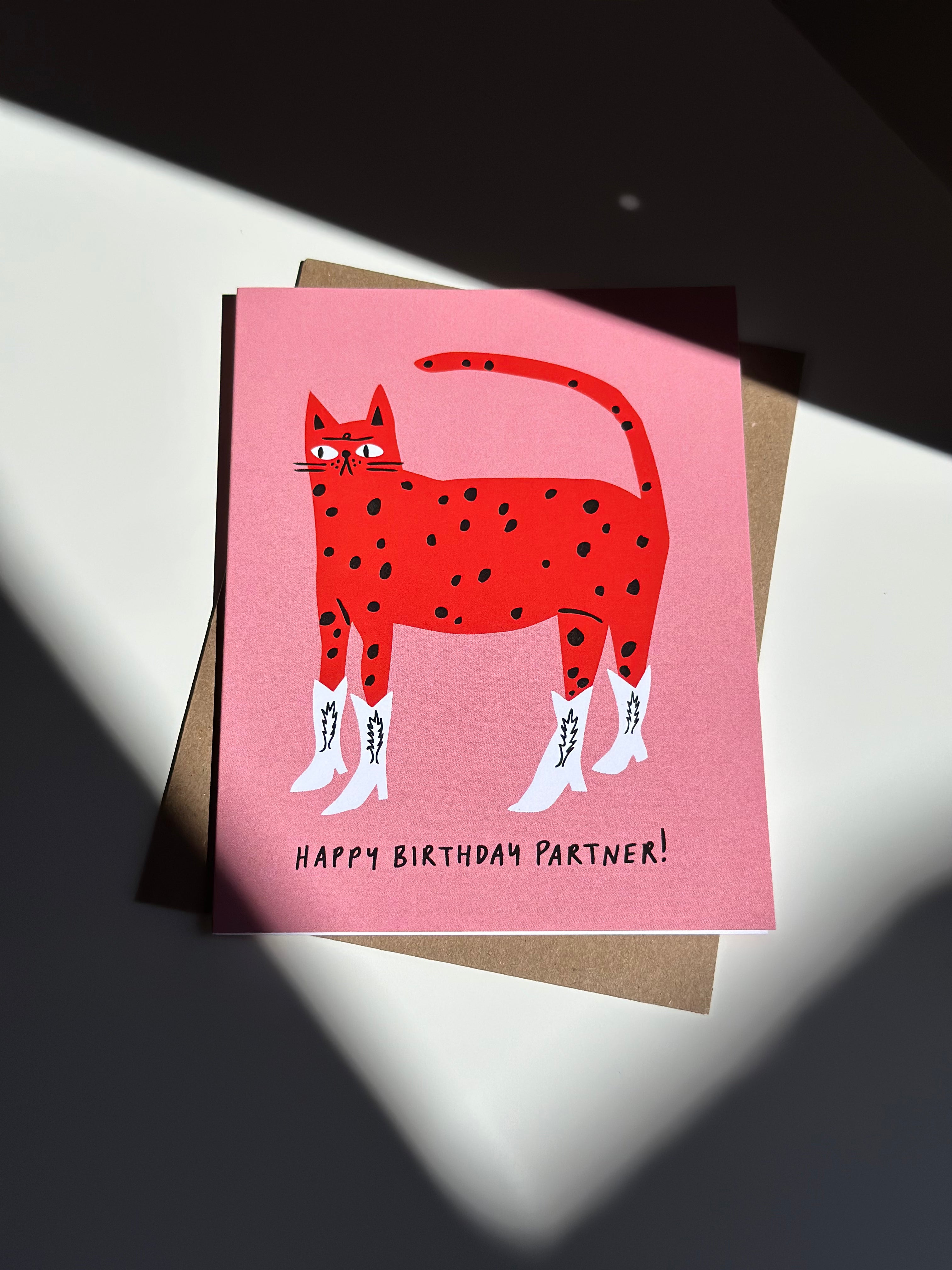 Western-themed cat birthday card with red spotted cat wearing white cowboy boots on pink background, captioned ‘Happy Birthday Partner!’ with brown kraft envelope.
