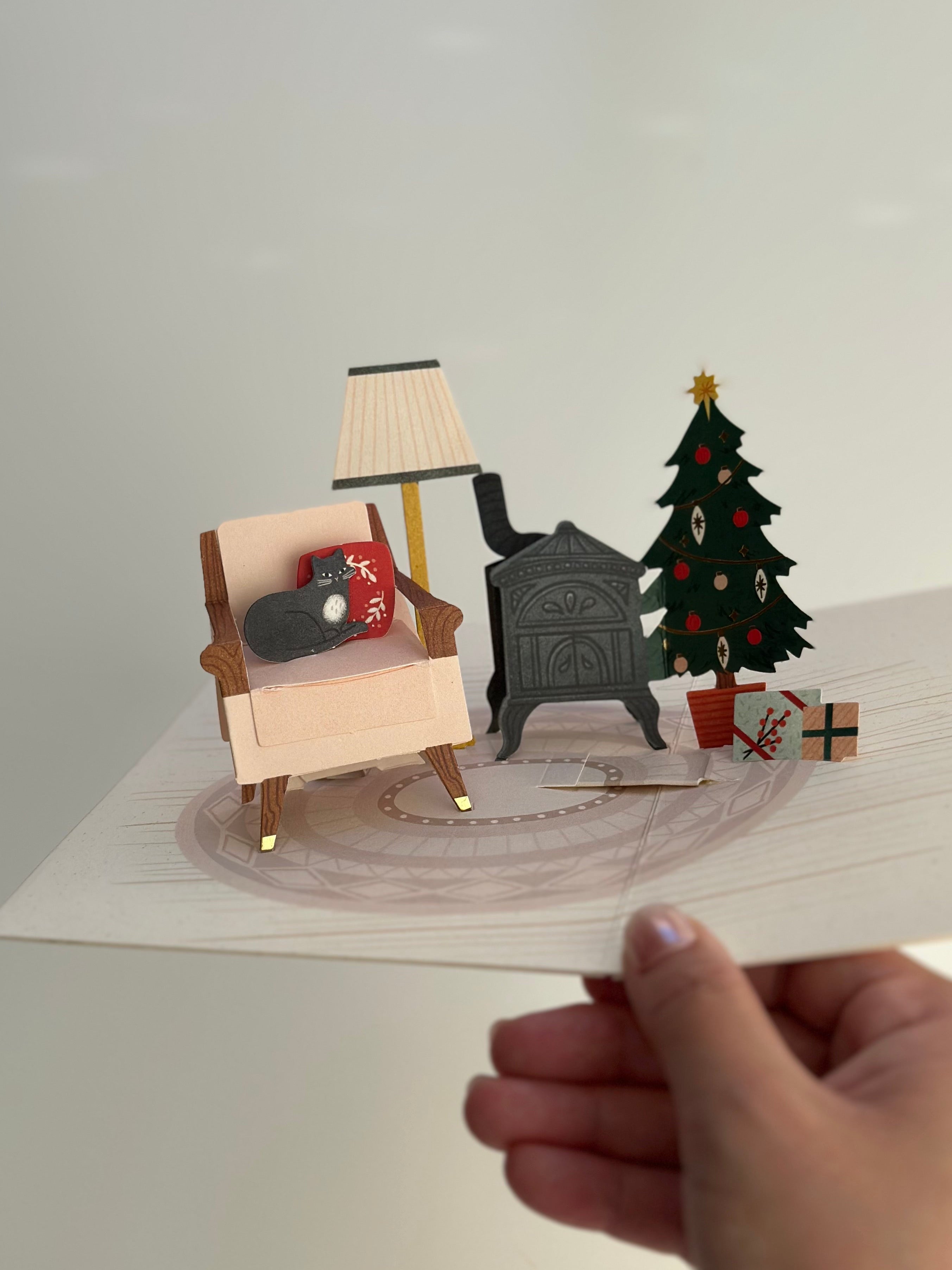 Freestanding pop-up Christmas card showing chair, lamp, wood stove, and tree with gifts.