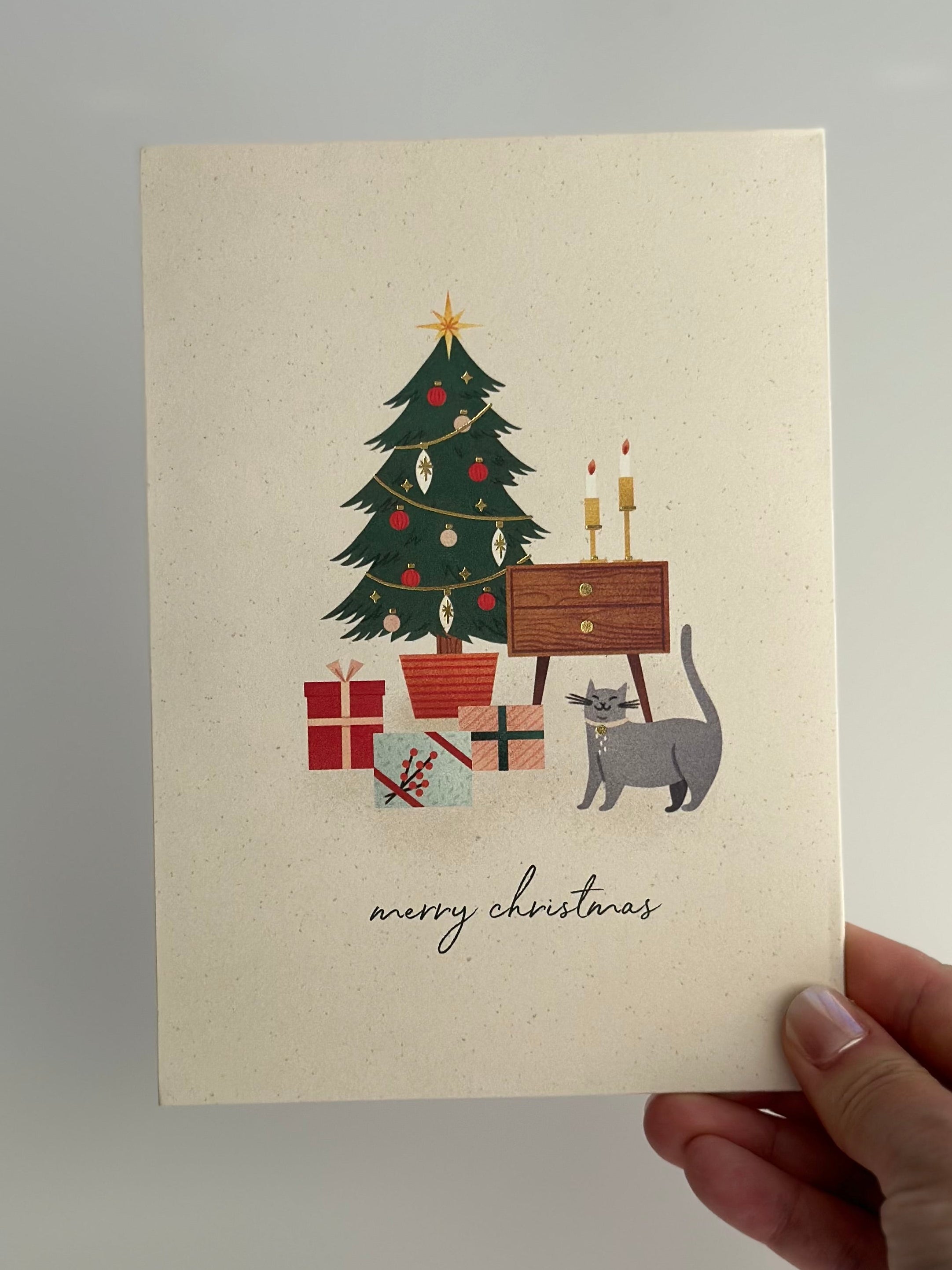 Hand holding a Merry Christmas card with tree, gifts, candles, and a gray cat illustration.