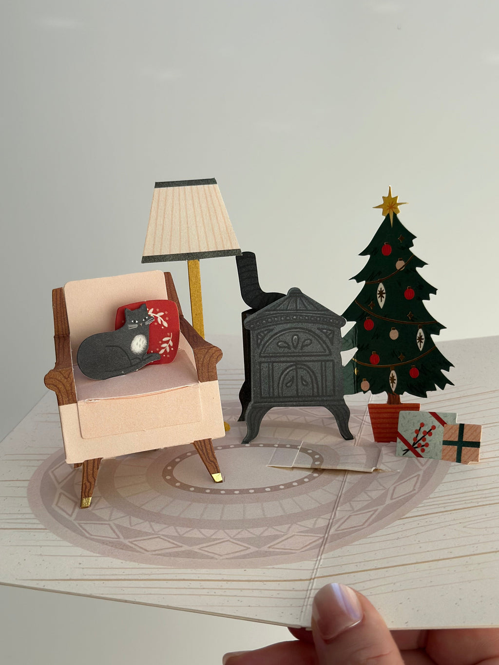 3D pop-up holiday room with armchair, sleeping cat, wood stove, and decorated Christmas tree.