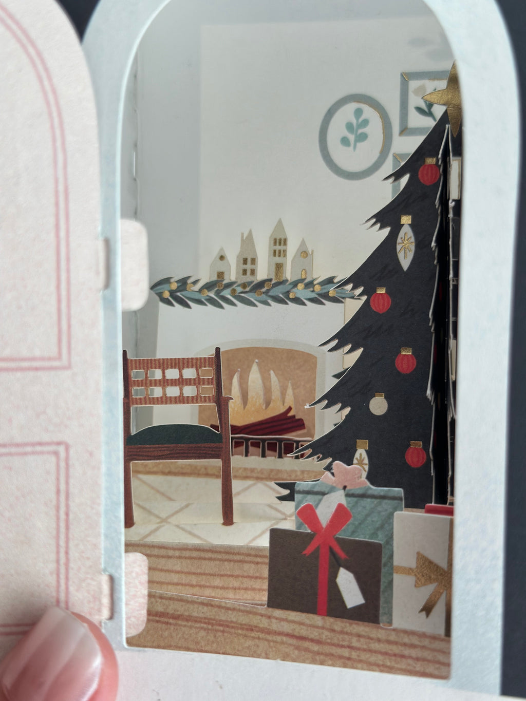 Hand opening wreath door on pop-up Christmas card to reveal cozy room scene.