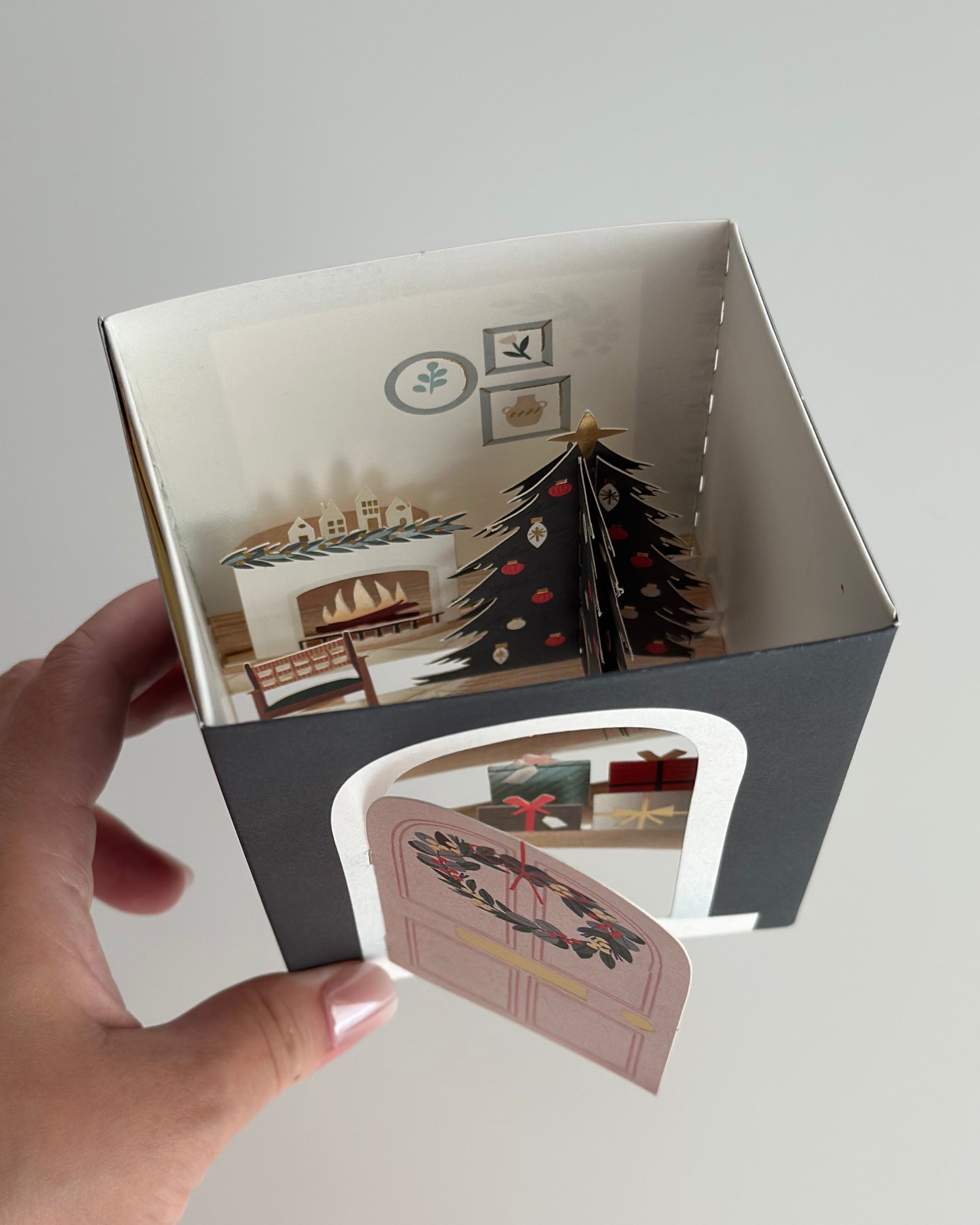 Hand holding pop-up Christmas card box showing door and cozy living room with tree and fireplace.