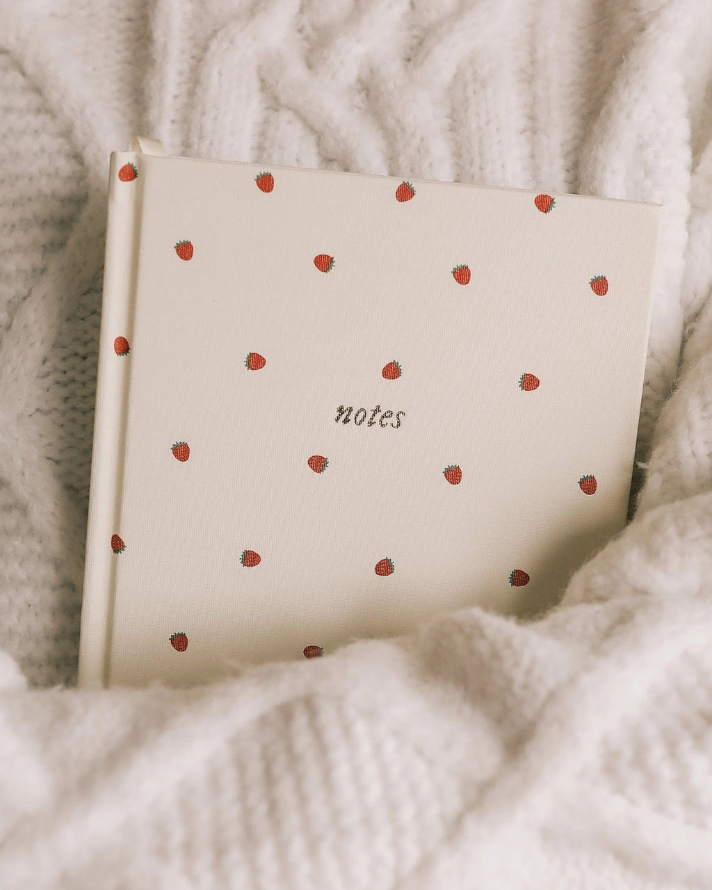 Strawberry linen A5 journal tucked into a soft knit blanket, cozy aesthetic notebook ideal for journaling, note-taking, and thoughtful gifting.