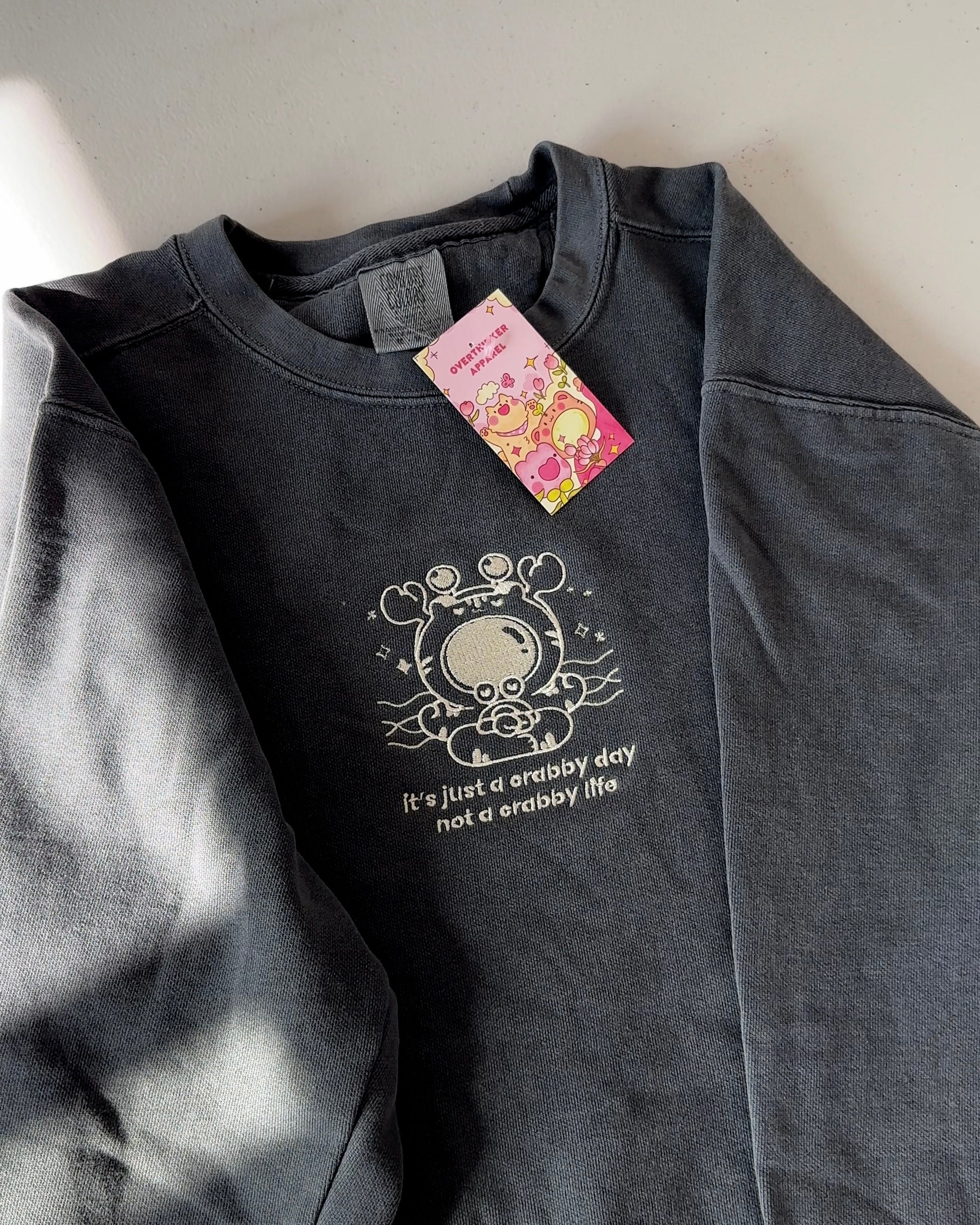 Charcoal crewneck sweatshirt laid flat featuring an embroidered crab illustration and the text “it’s just a crabby day, not a crabby life.”