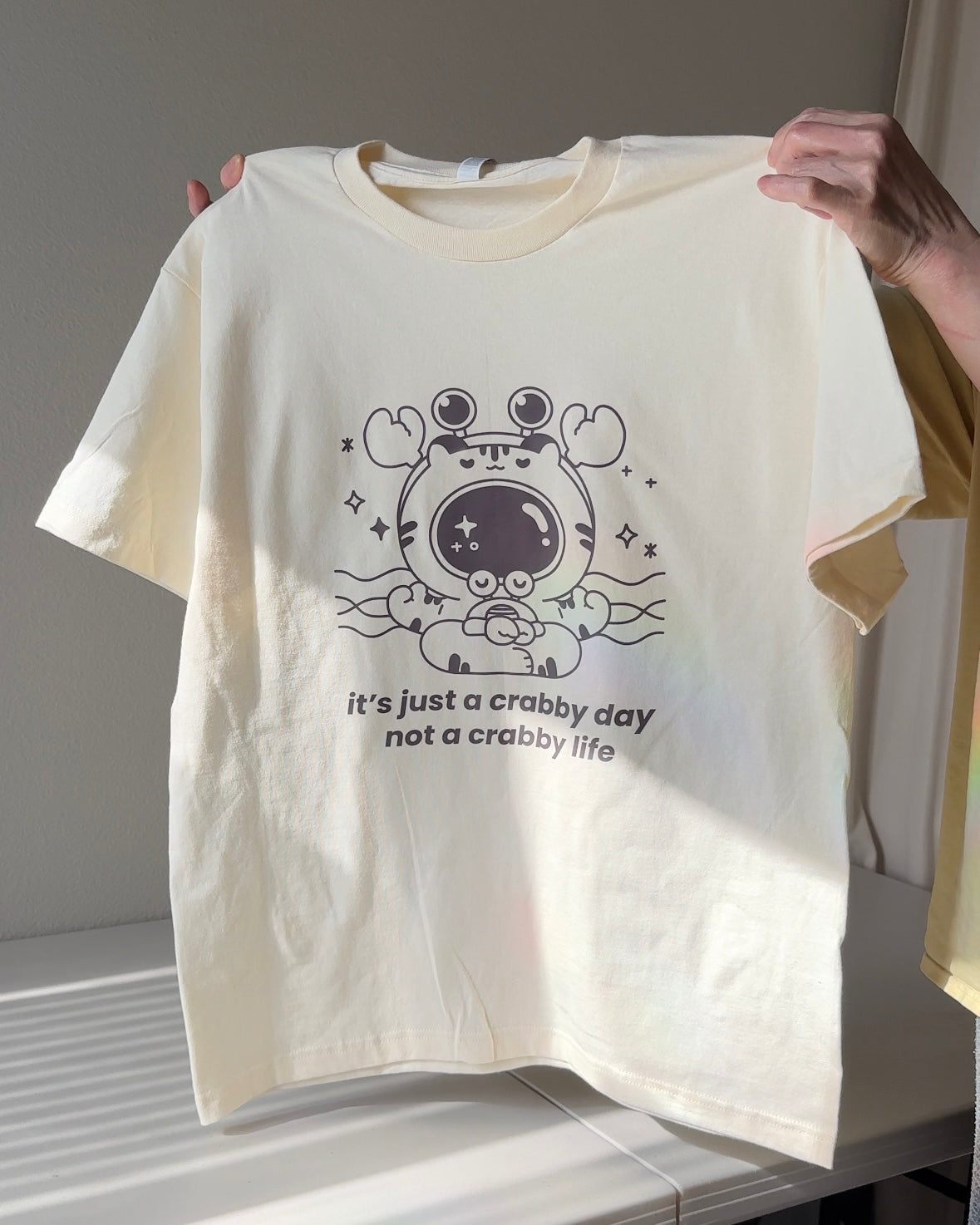 Ivory graphic t-shirt featuring a cute meditating crab with the phrase “it’s just a crabby day not a crabby life,” a mental health humor tee made for comfort and perspective
