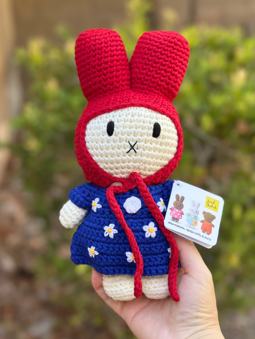 Handmade crochet bunny doll wearing a red hood and blue dress with white daisy flowers, part of the Just Dutch Miffy collection, shown with brown kraft tag.