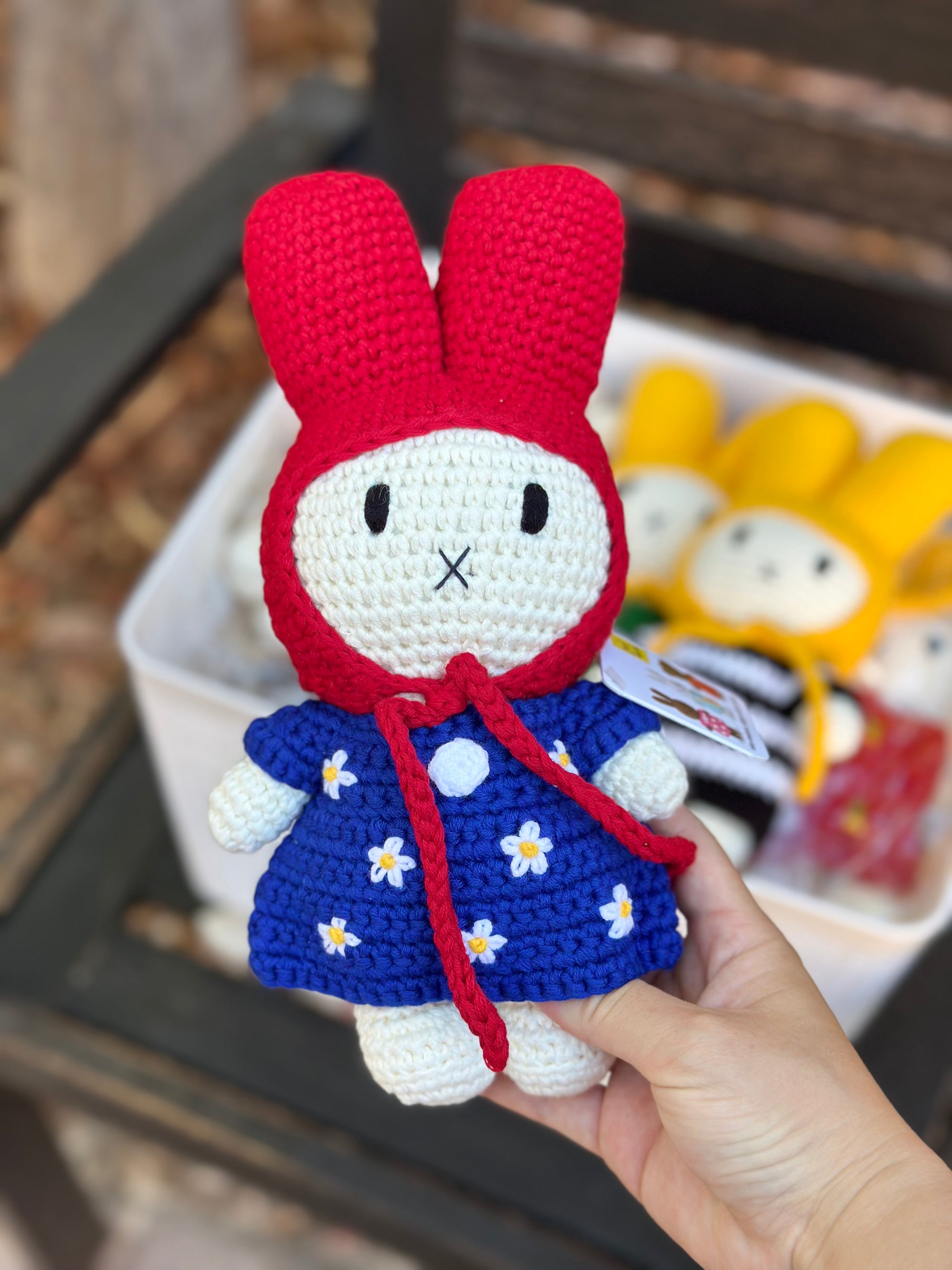 Handmade crochet bunny doll wearing a red hood and blue dress with white daisy flowers, part of the Just Dutch Miffy collection, shown with brown kraft tag.