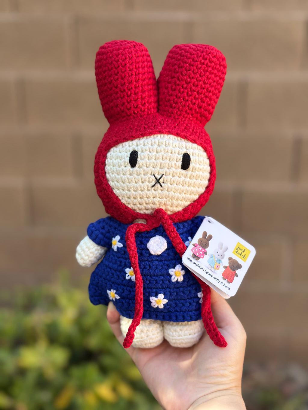Handmade crochet bunny doll wearing a red hood and blue dress with white daisy flowers, part of the Just Dutch Miffy collection, shown with brown kraft tag.