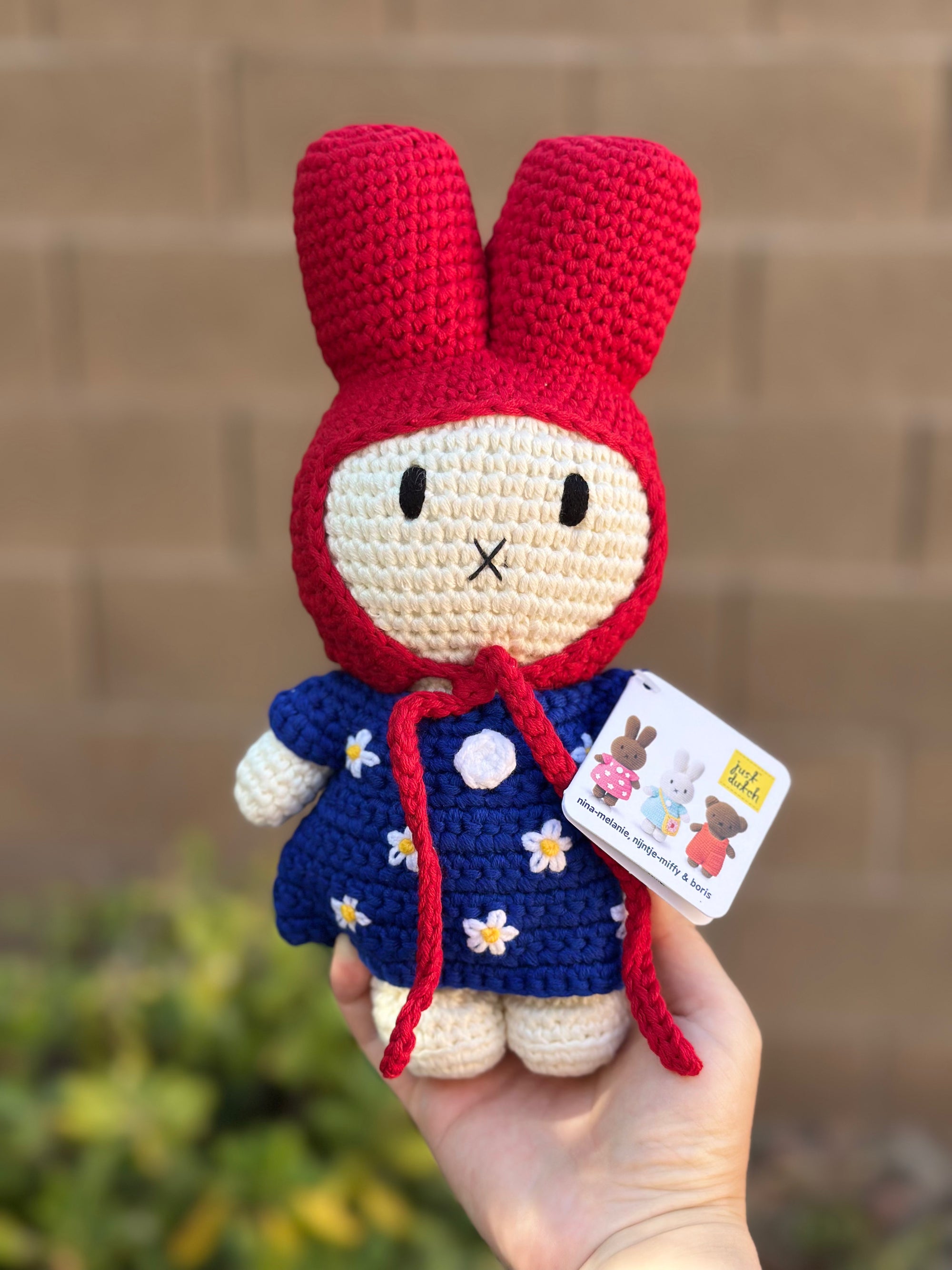 Handmade crochet bunny doll wearing a red hood and blue dress with white daisy flowers, part of the Just Dutch Miffy collection, shown with brown kraft tag.