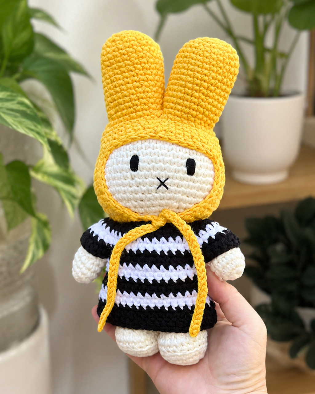 A handmade crocheted amigurumi bunny doll with yellow ears, a black and white striped outfit, and a yellow drawstring, held in a person's hand against a blurred indoor background with green plants.