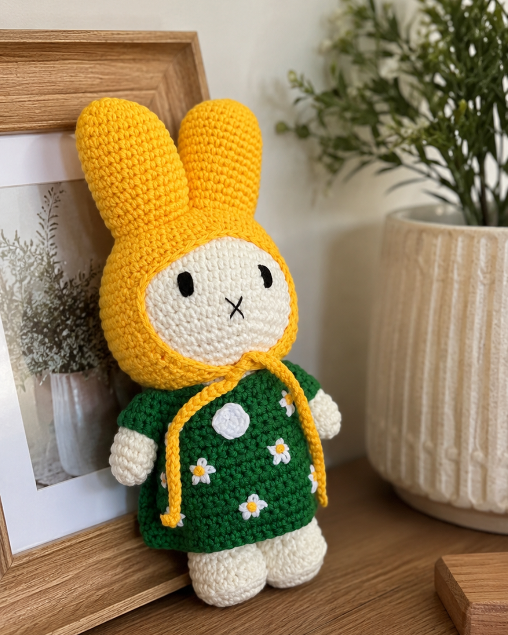 A crocheted bunny doll with a yellow hood, white face and legs, and a green dress decorated with small white flowers, standing on a wooden surface next to a framed picture and a potted plant.