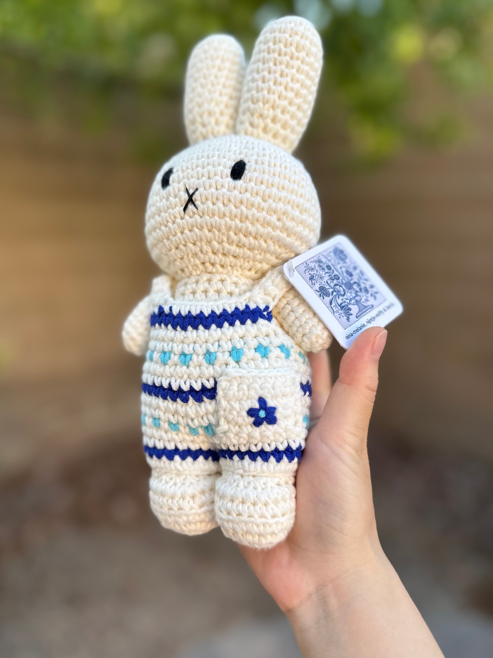 Handmade crochet Miffy plush in Delft blue floral jumpsuit with pocket detail, held outdoors in natural light.