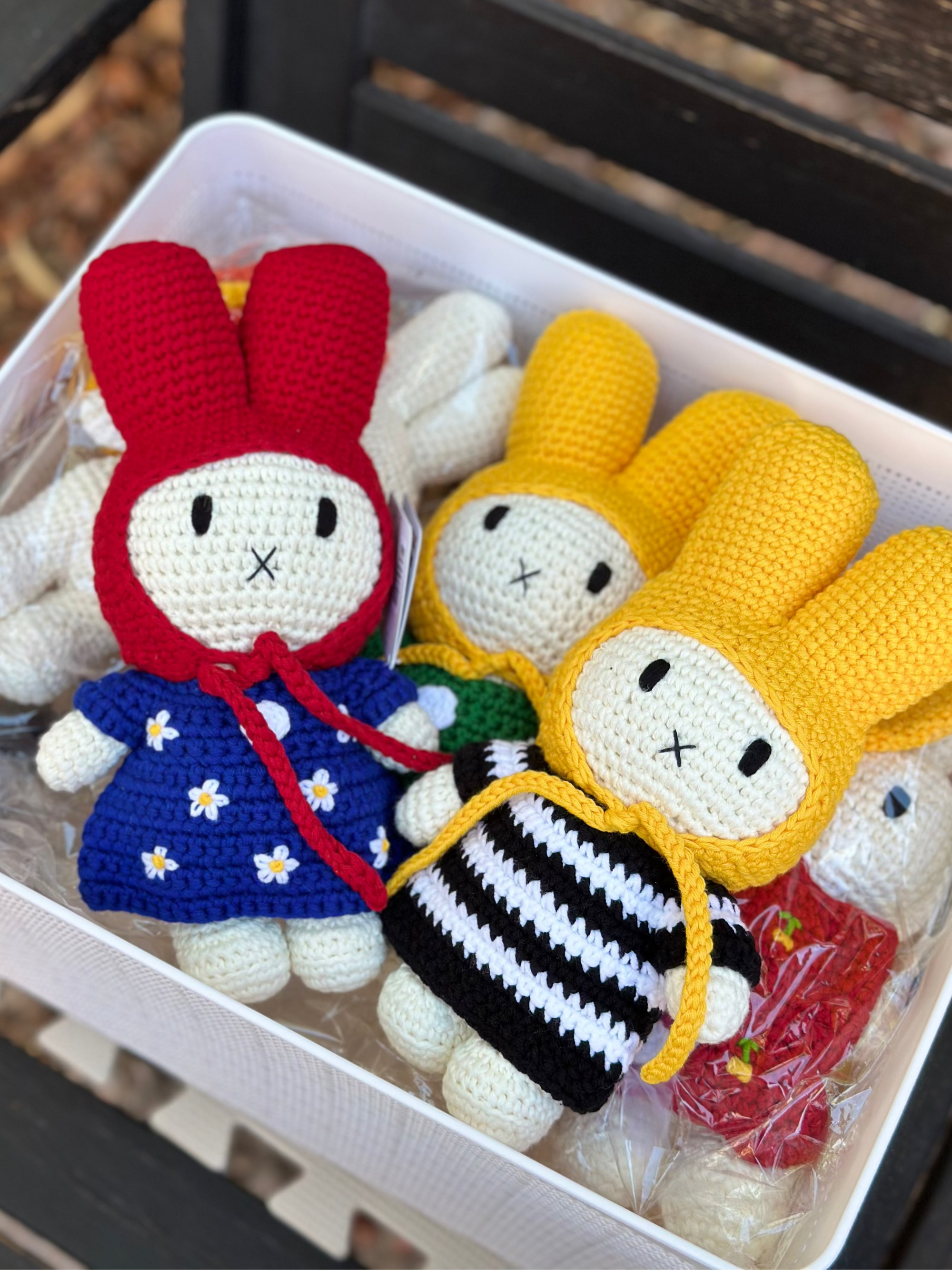 Box of handmade crochet Miffy dolls, including red-hooded Miffy in a blue daisy dress and yellow-hooded Miffy in black-and-white striped dress, part of the Just Dutch Miffy collection.