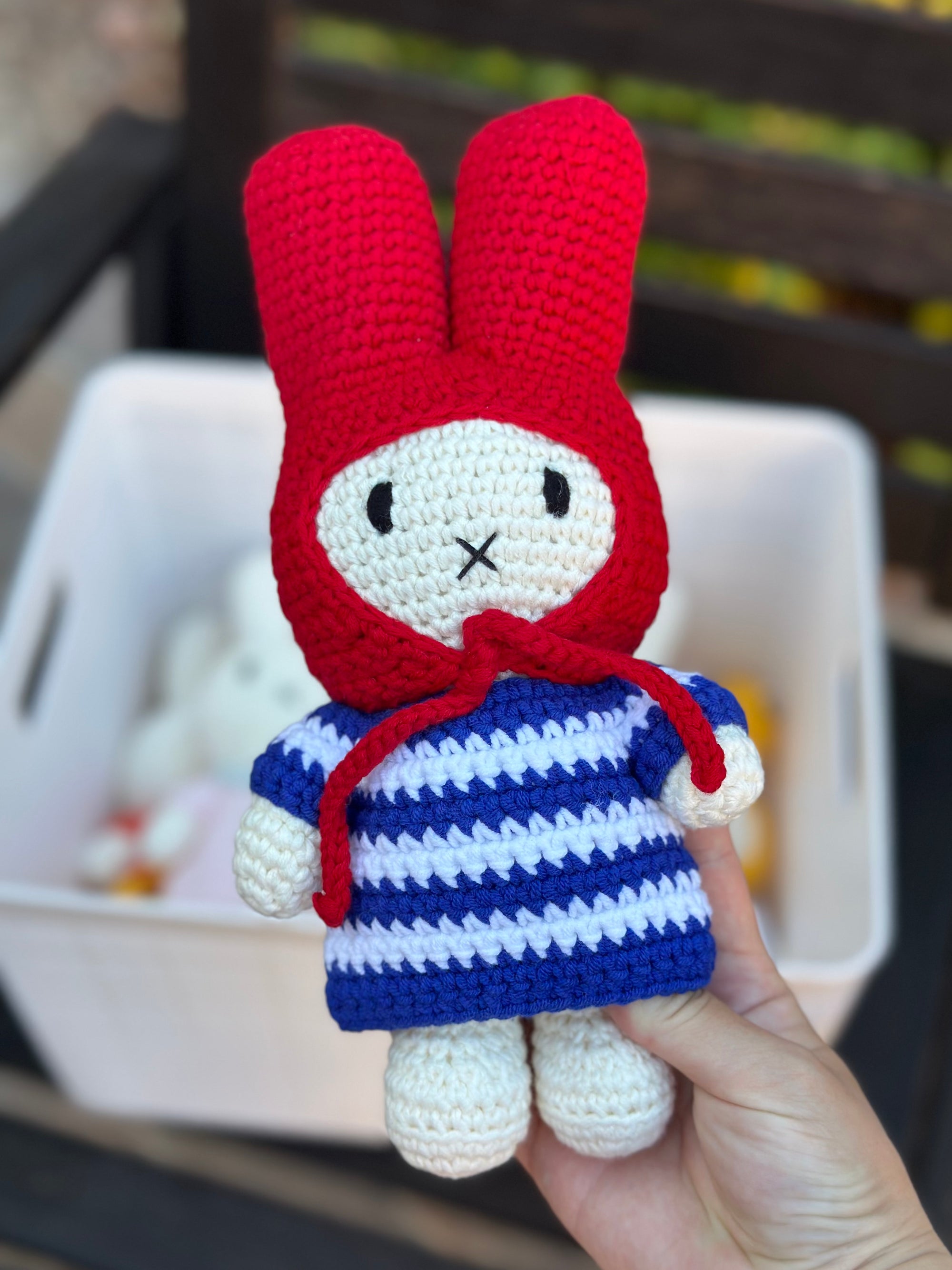 Handmade crochet Miffy plush in striped dress and red bonnet, held in front of a storage basket with other crochet dolls inside.