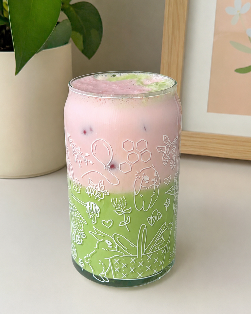 A frosted glass can-shaped tumbler with white line-art illustrations of bunnies, flowers, bees, and hearts, filled with a pink and green layered drink and served with ice, set on a tabletop next to a potted plant.