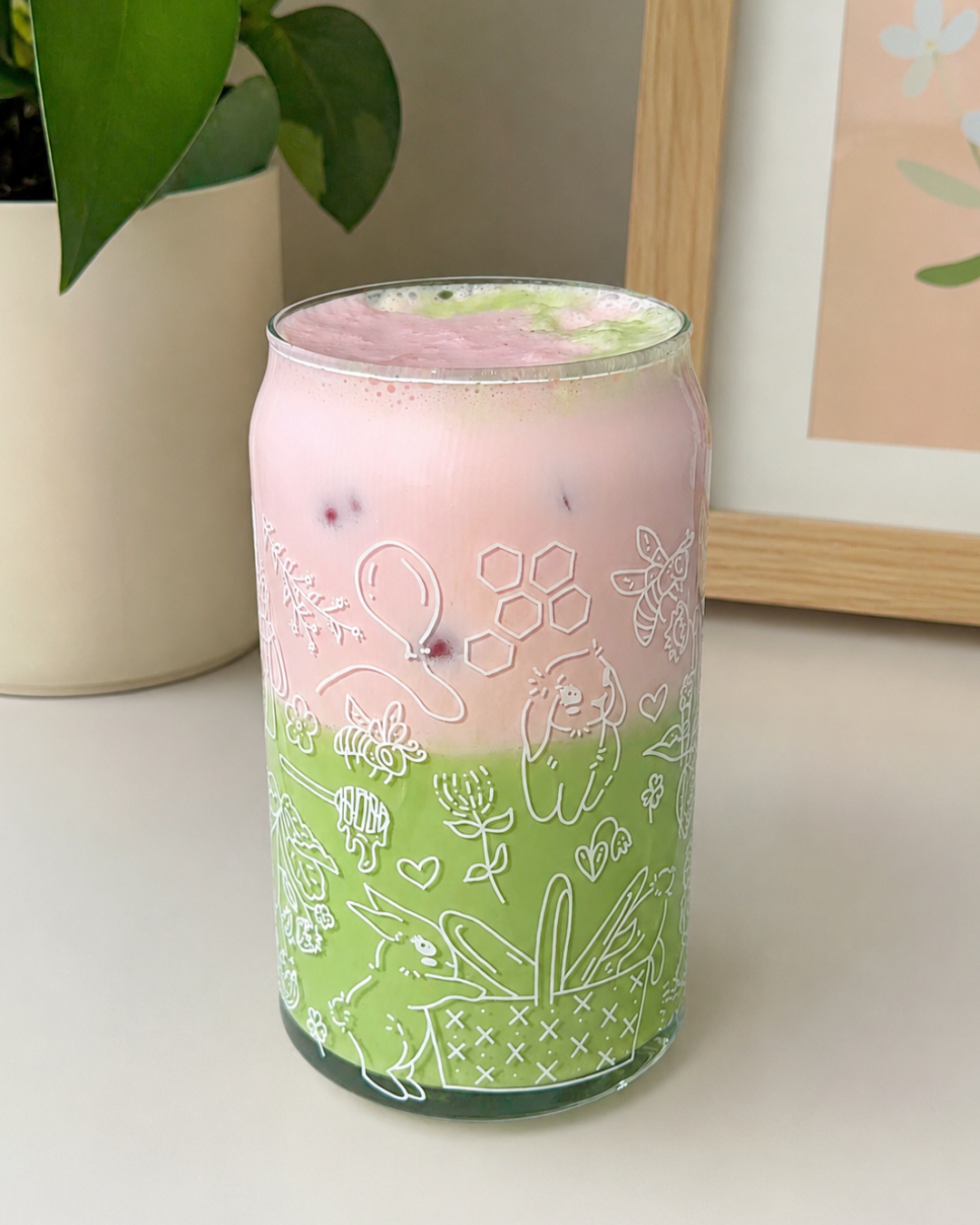 A frosted glass can-shaped tumbler with white line-art illustrations of bunnies, flowers, bees, and hearts, filled with a pink and green layered drink and served with ice, set on a tabletop next to a potted plant.