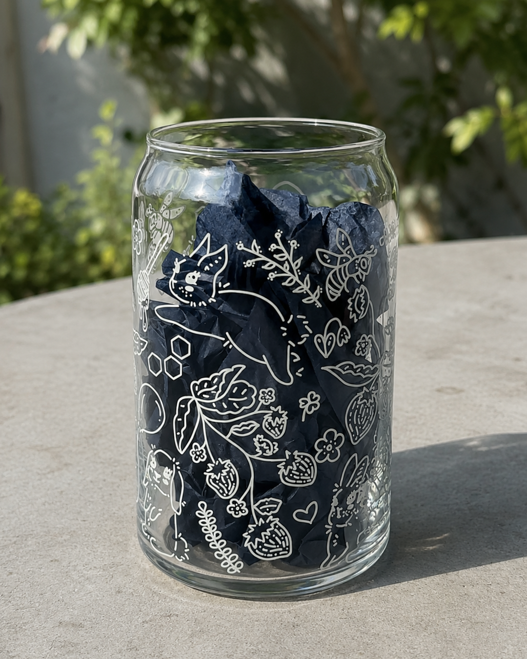 A clear glass jar filled with dark navy blue bandanas printed with white botanical and floral line drawings, sitting on a concrete surface outdoors with blurred green foliage in the background.