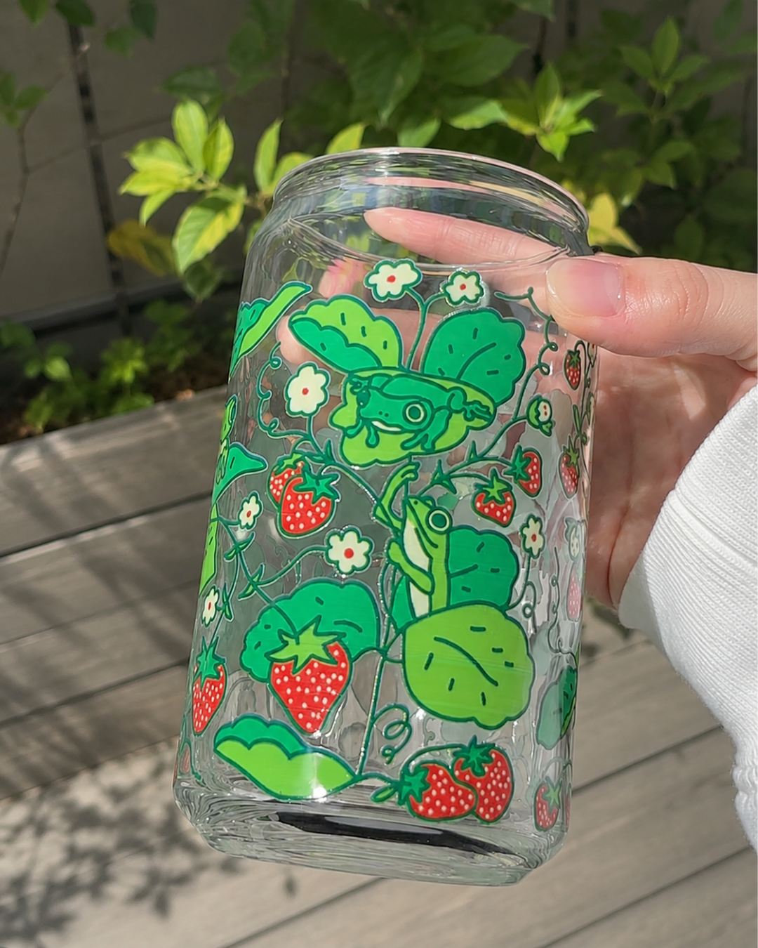 A hand holding a clear glass jar decorated with a green strawberry and frogs pattern, set against a background of a wooden deck and green foliage.