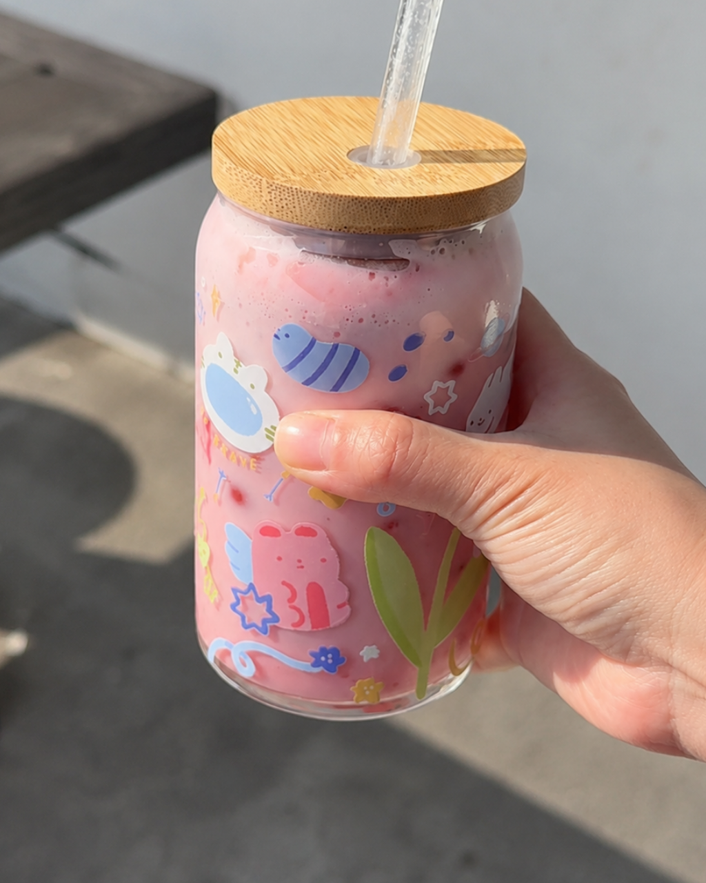 A hand holding a clear glass straw cup with a light wood lid, filled with a pink drink. The cup is decorated with colorful cartoon illustrations, including a pink character, blue stars, and green leaves.