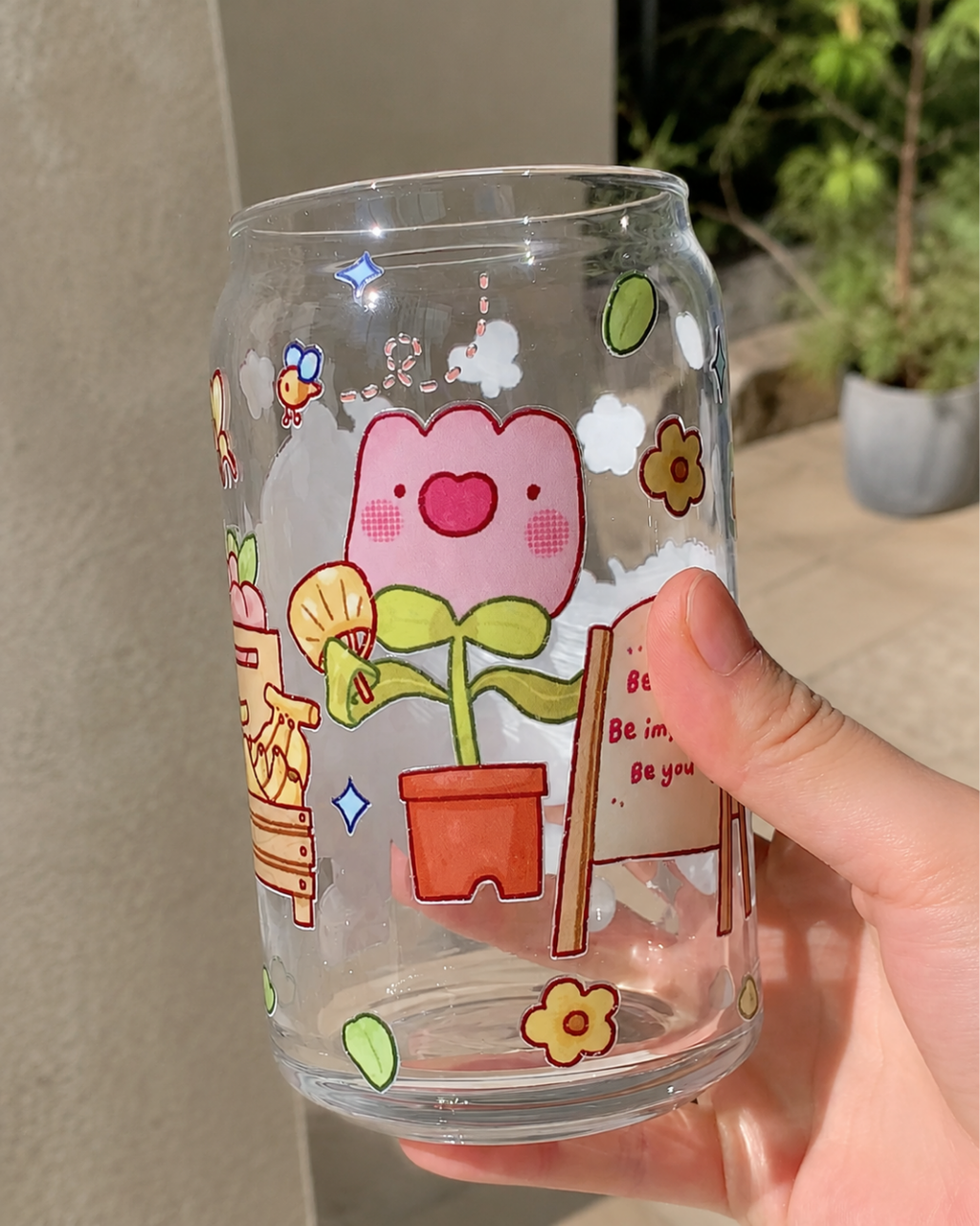 A hand holding a clear glass can-shaped tumbler decorated with cute cartoon illustrations, including a smiling pink flower in an orange pot, small flowers, stars, and a sign that reads 'Be brave be imperfect Be you'.