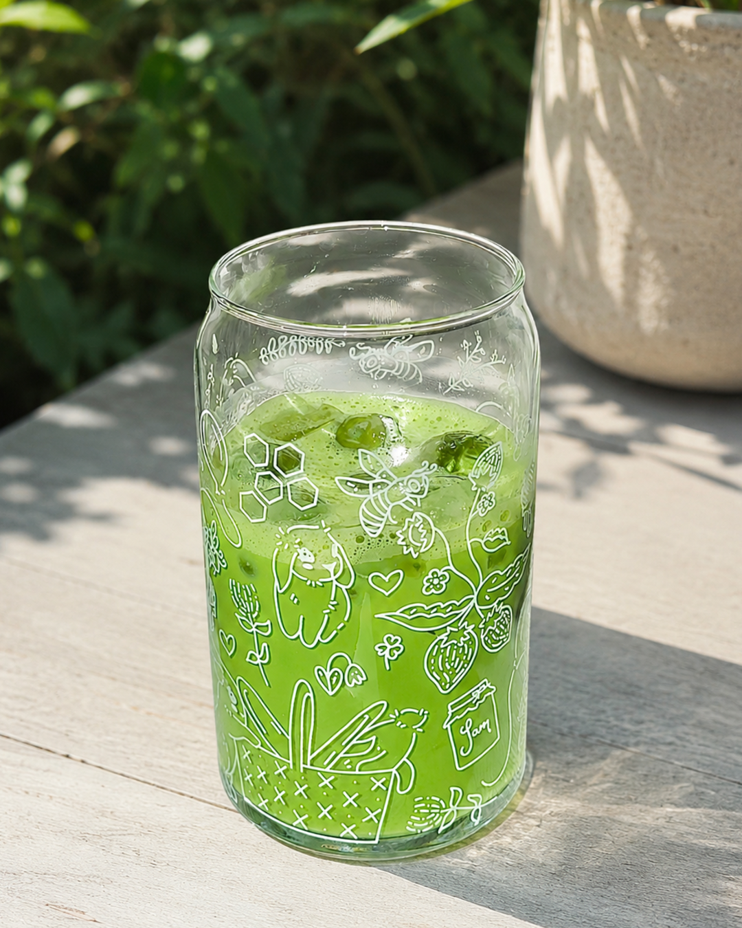A clear glass jar tumbler with white hand-drawn bee and strawberry illustrations, filled with a bright green drink and placed on a light wooden table outdoors.