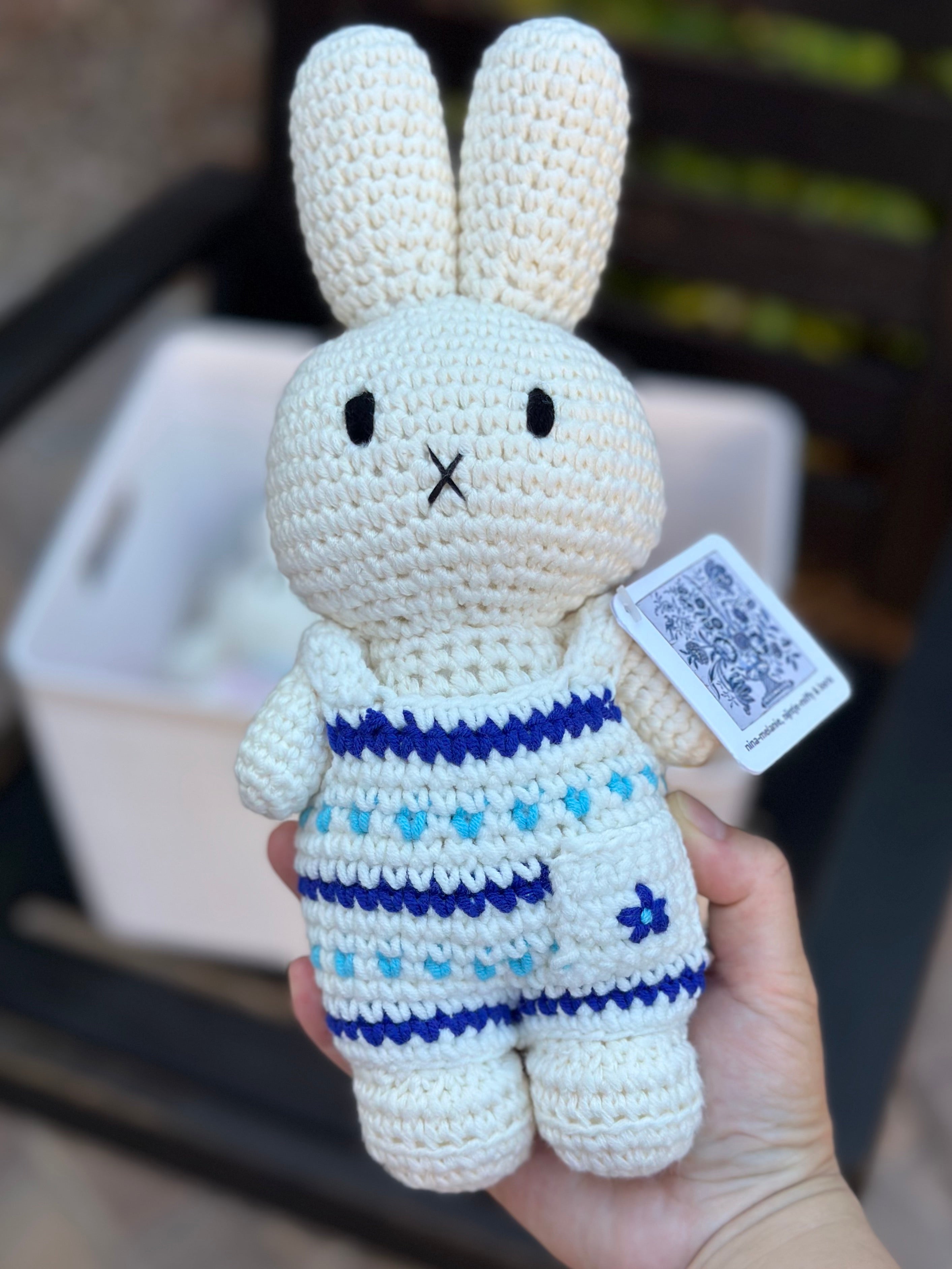 Delft blue crochet Miffy plush in floral jumpsuit held in front of a basket of other handmade dolls.