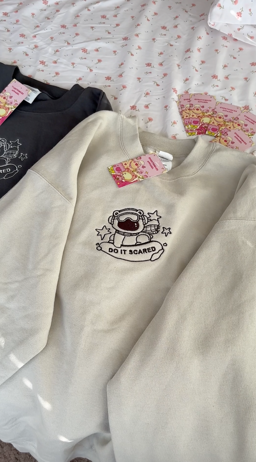 Flat lay of beige and charcoal gray oversized embroidered sweatshirts featuring a polar bear and “Do It Scared” message, showing both neutral color options.