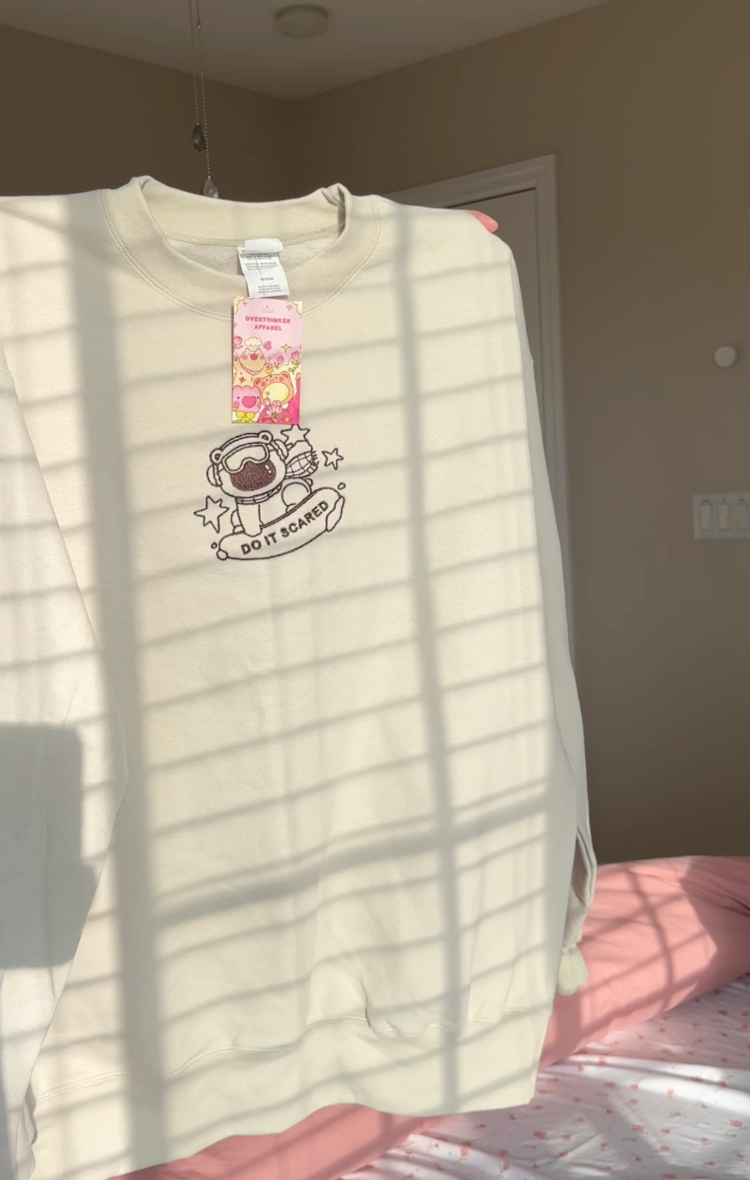 Beige oversized crewneck sweatshirt with subtle “Do It Scared” polar bear embroidery, hanging in natural window light for a cozy, calming look.