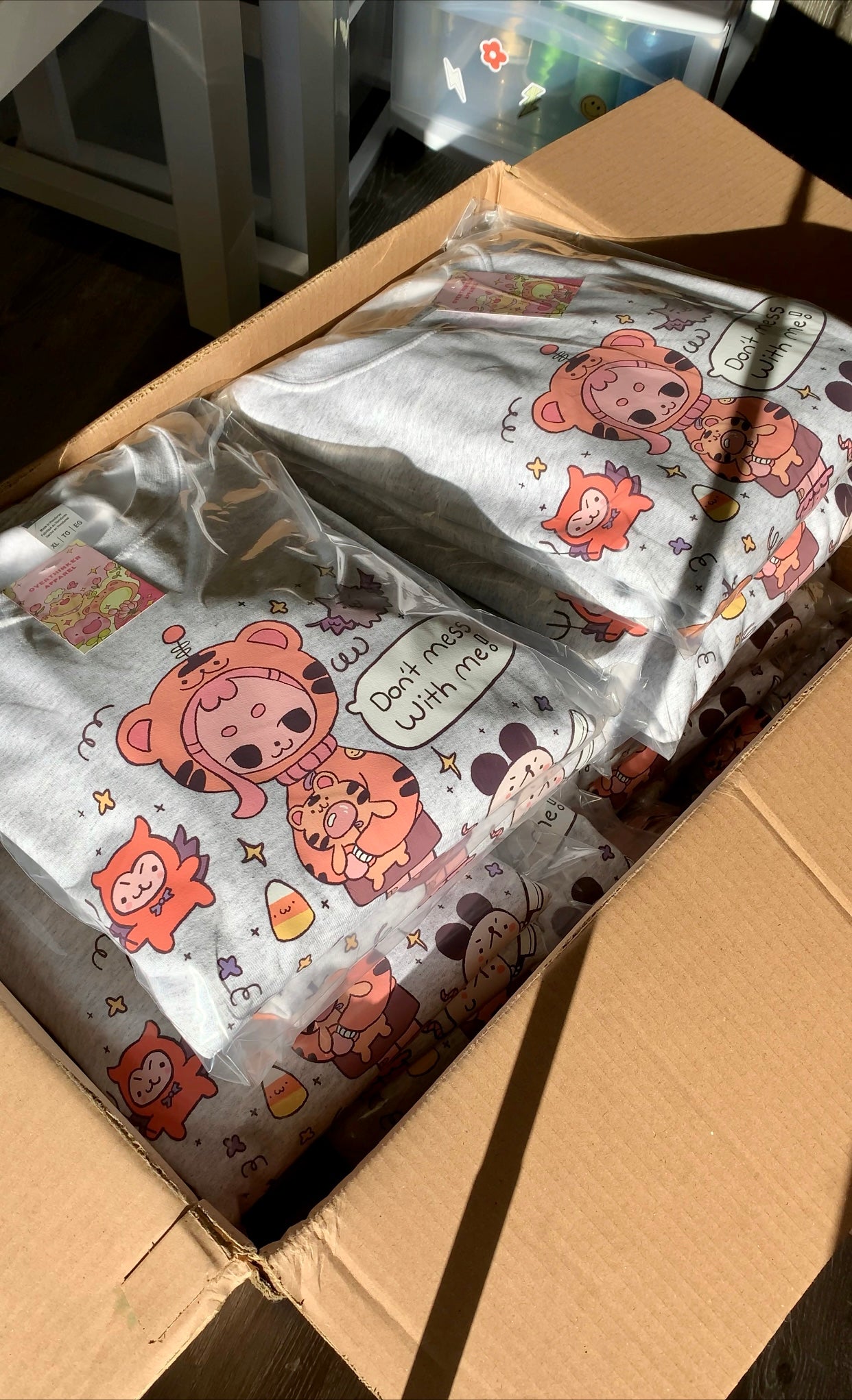 Packaged ash gray graphic sweatshirts stacked in a box, featuring a cute illustrated character and the phrase “Don’t mess with me,” prepared in small batches for shipping.