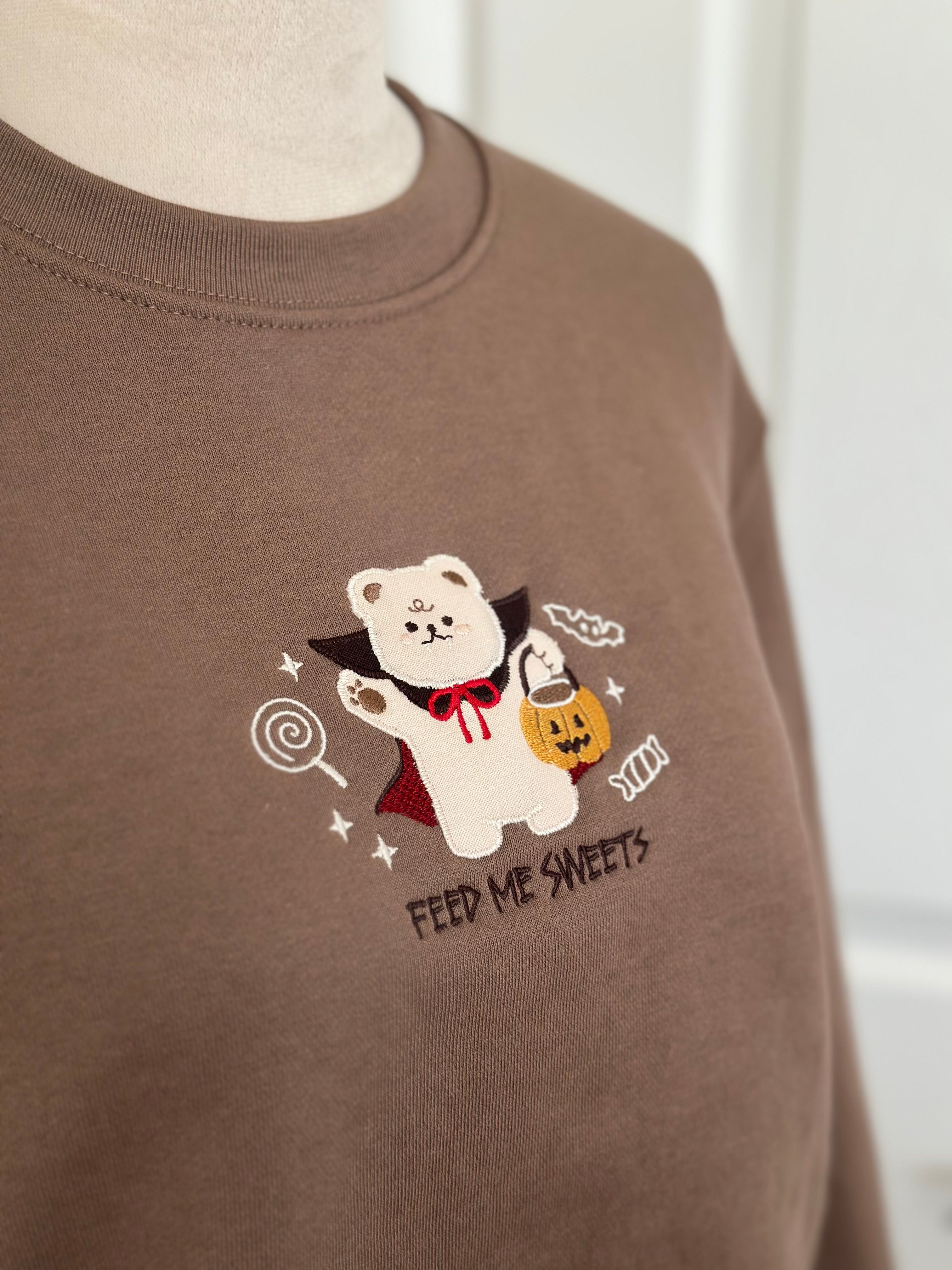 Embroidered detail of vampire bear holding pumpkin candy bucket on brown sweatshirt with “Feed Me Sweets” text.