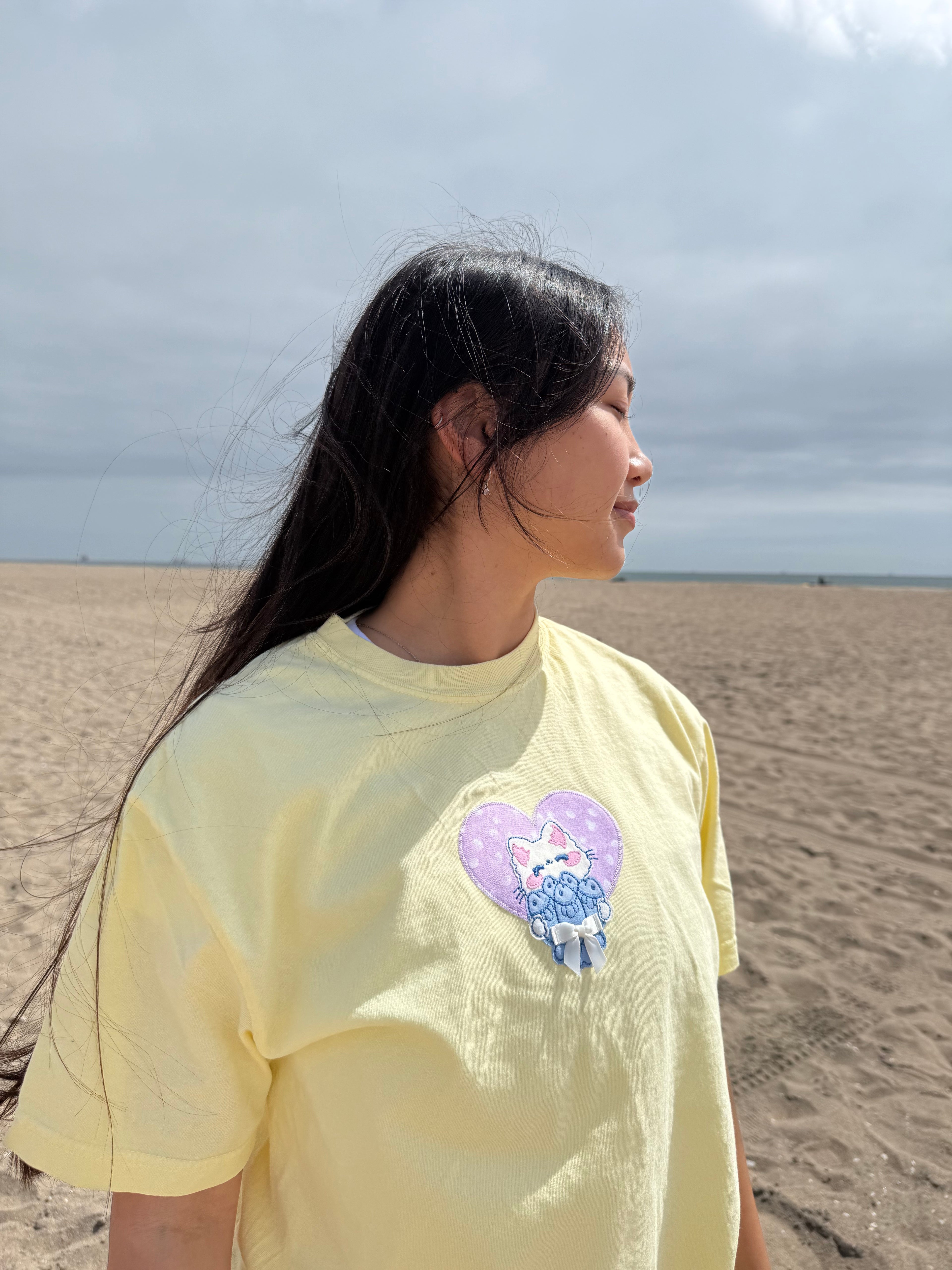 Close-up side view of embroidered cat patch on pastel yellow t-shirt, showing lilac heart background and soft handmade stitching.
