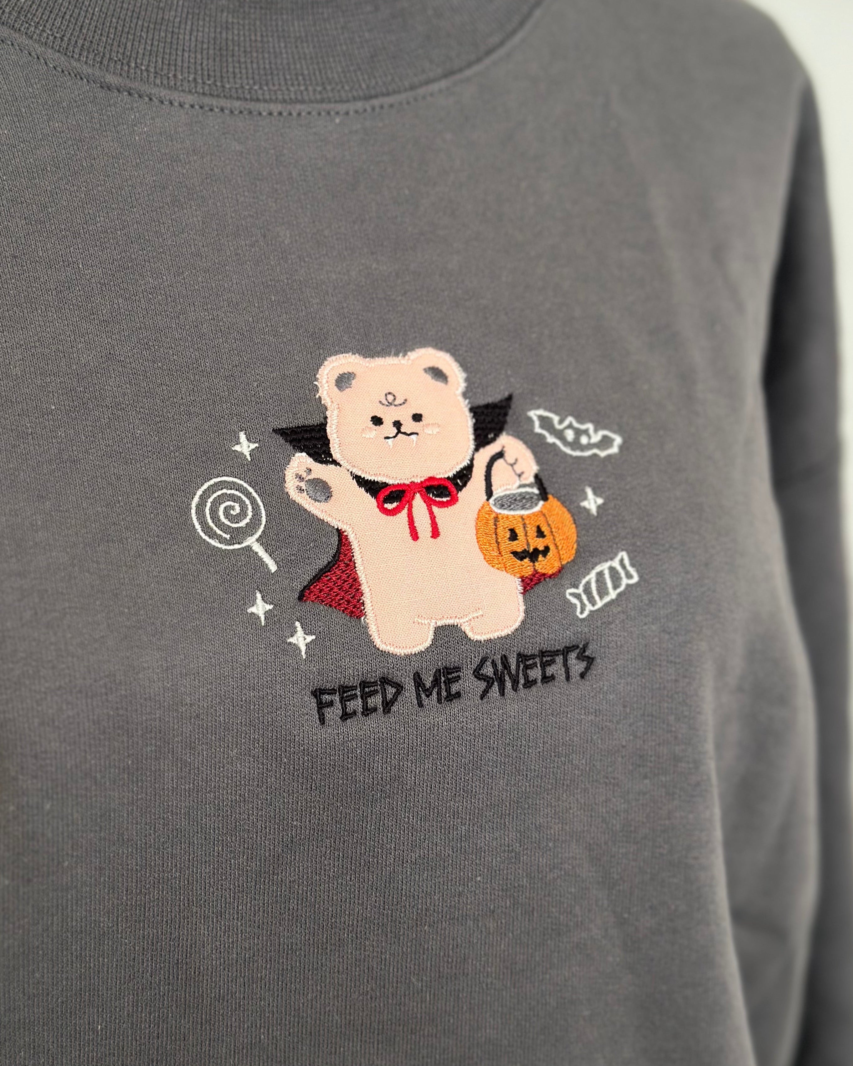 Embroidered detail of a bear in a vampire costume holding a pumpkin candy bucket on a gray sweatshirt, with “Feed Me Sweets” text.