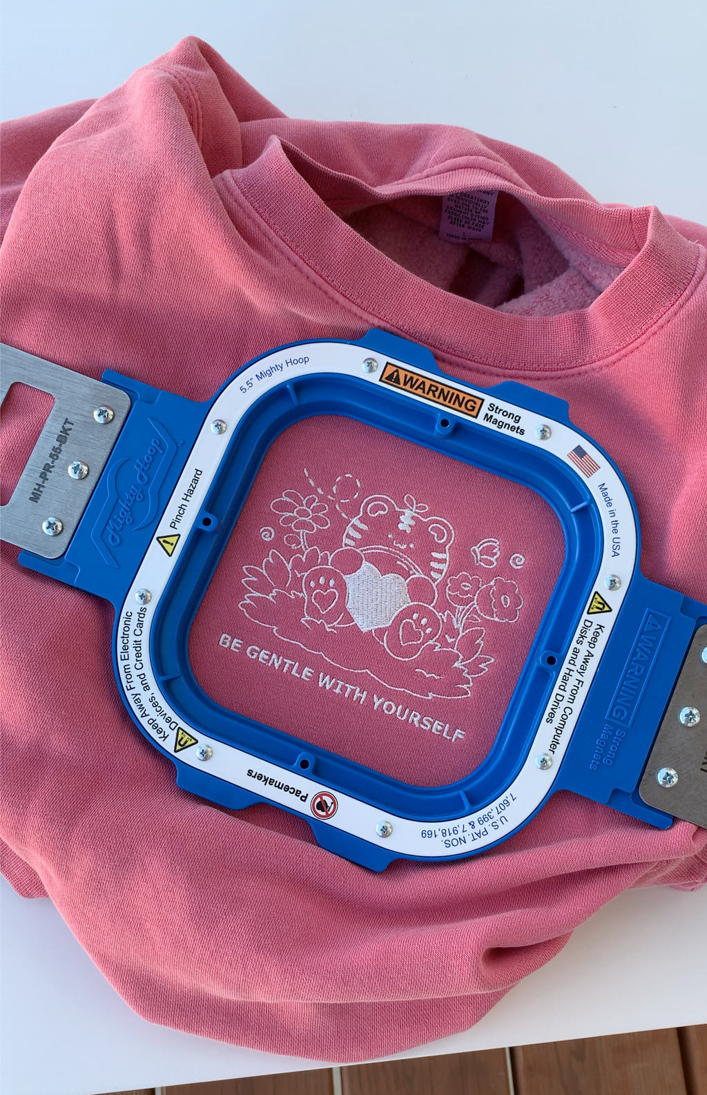 In-progress embroidery of a tiger holding a heart on a rose pink sweatshirt with the phrase “Be Gentle With Yourself”