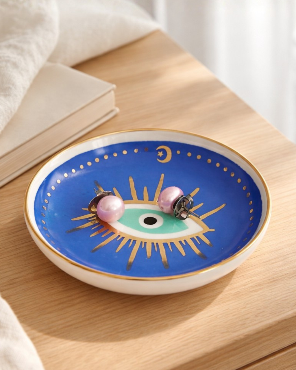 Blue evil eye jewelry dish with pearl earrings on wooden bedside table next to book, decorative ring holder for nightstand jewelry storage