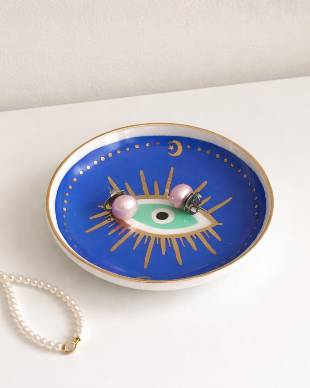 Blue evil eye ring holder dish with pearl earrings and pearl necklace on minimal white surface, jewelry organizer tray for vanity decor