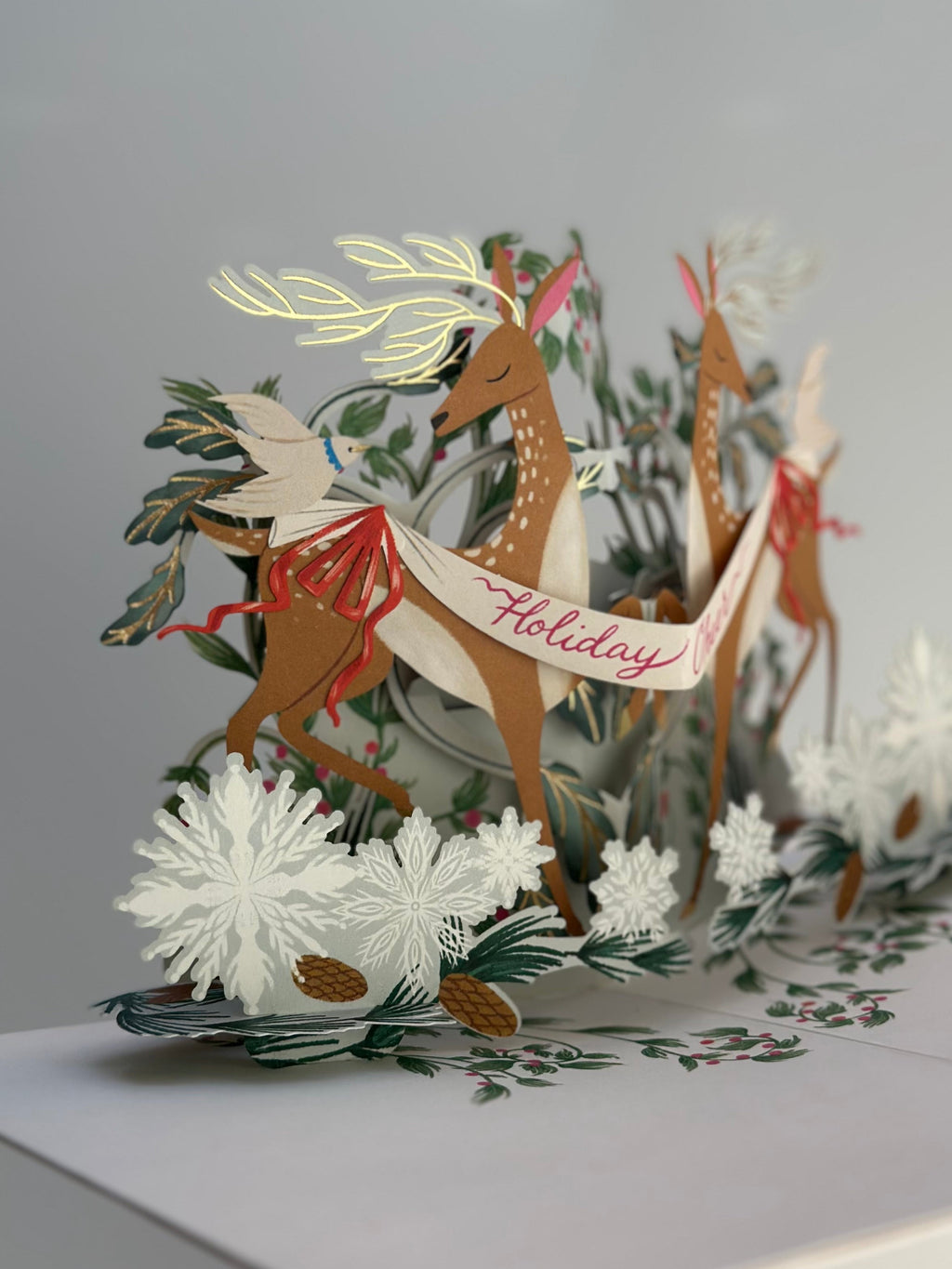 3D pop-up Christmas card showing graceful deer, birds, pine branches, and snowflakes.