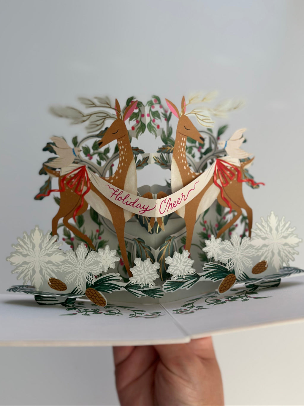 Center view of festive deer pop-up Christmas card surrounded by snowflakes and evergreens.