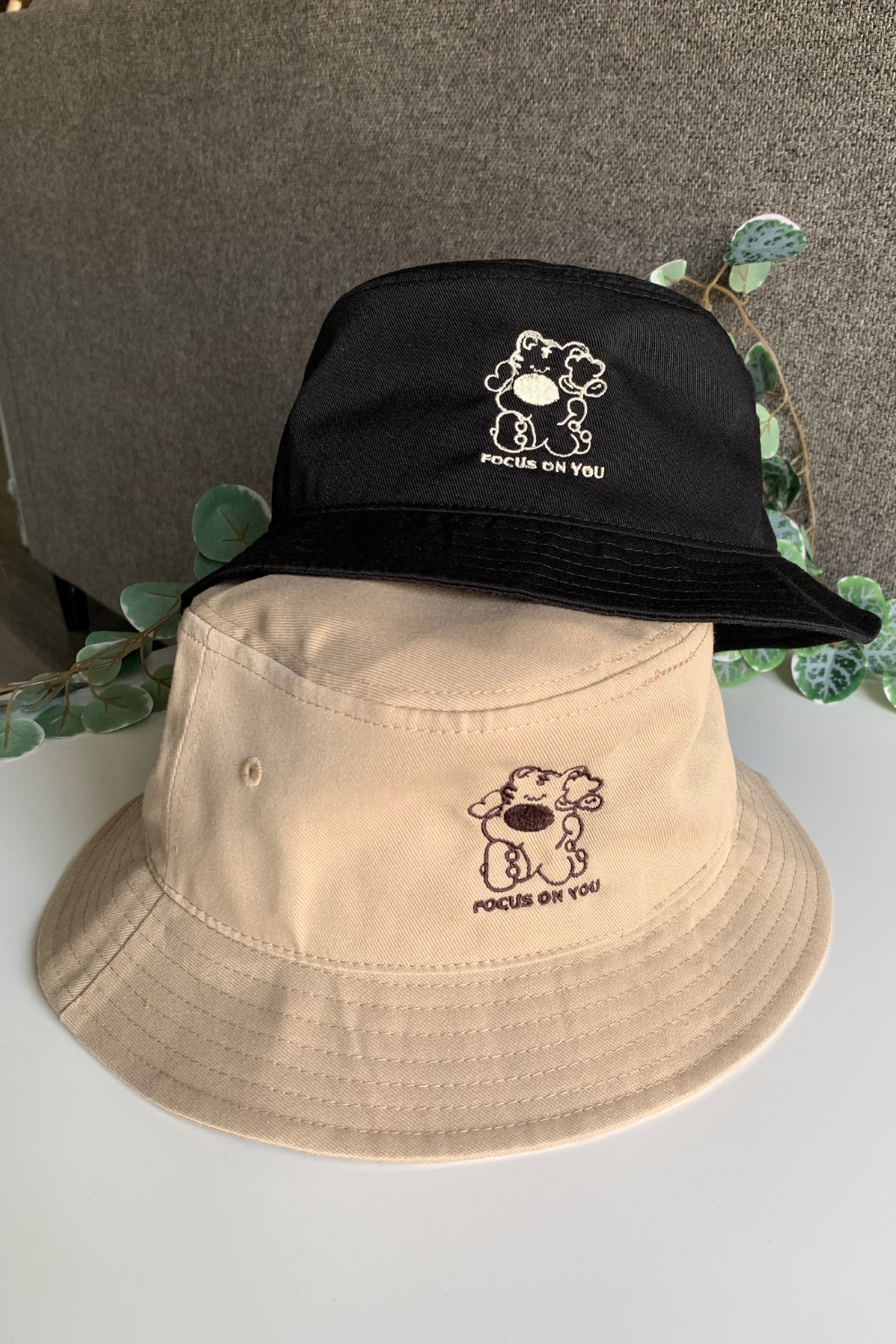 Tan bucket hat with embroidered bear character and "Focus on You" text, stacked beneath a black bucket hat with the same design, against a neutral background with greenery.
