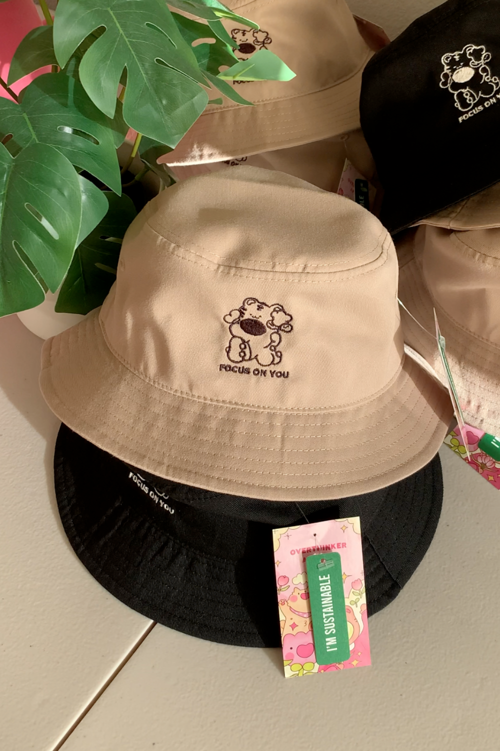 Tan bucket hat with embroidered bear character and "Focus on You" text, styled next to a green monstera plant.
