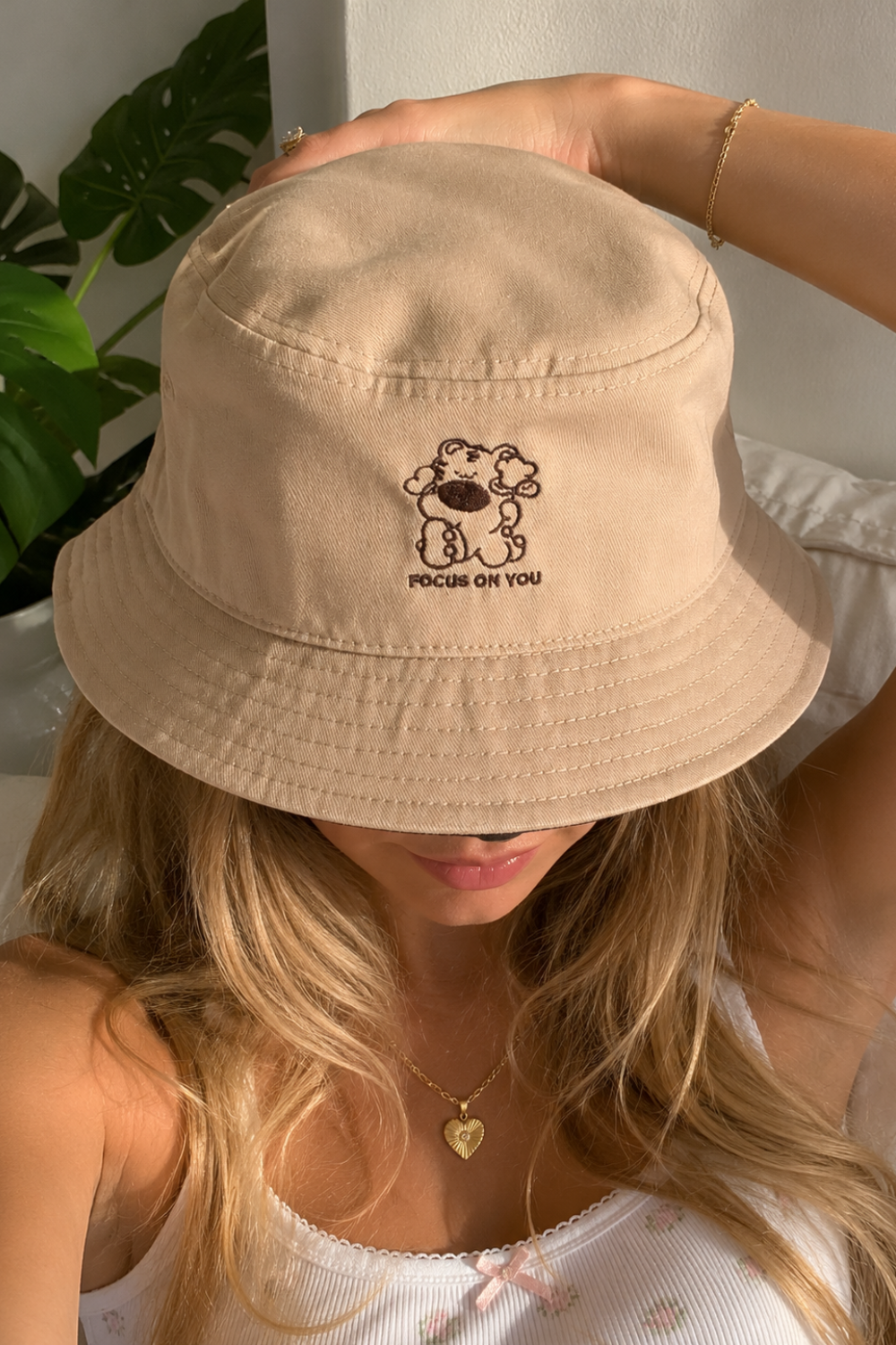 Woman wearing a tan bucket hat with embroidered bear character and "Focus on You" text, close-up shot with a heart pendant necklace and a monstera plant in the background.
