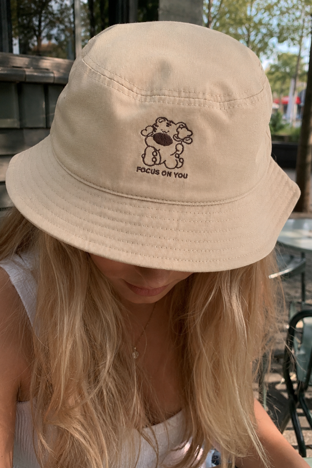 Woman wearing a tan bucket hat with embroidered bear character and "Focus on You" text, outdoors near a cafe with trees in the background.
