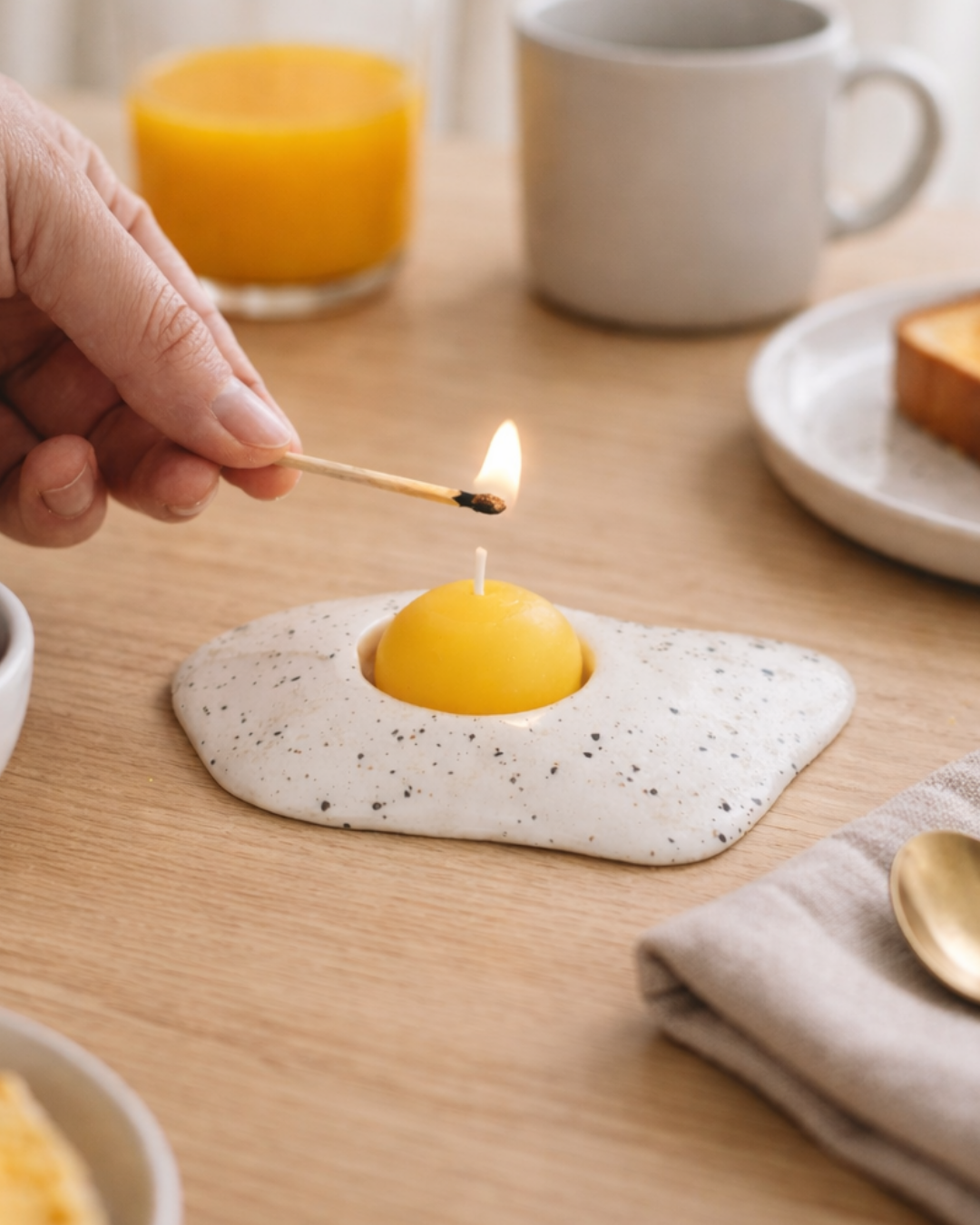 Lighting a fried egg candle on wooden breakfast table with coffee and toast, whimsical food shaped candle for quirky kitchen decor.