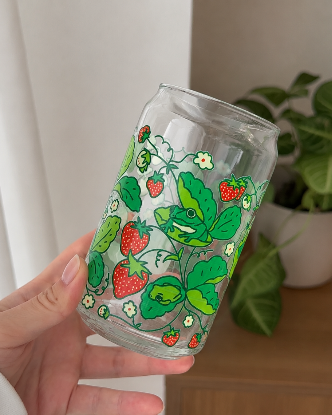 A hand holding a clear glass can-shaped tumbler decorated with a green strawberry and frogs pattern, with a blurred indoor plant in the background.