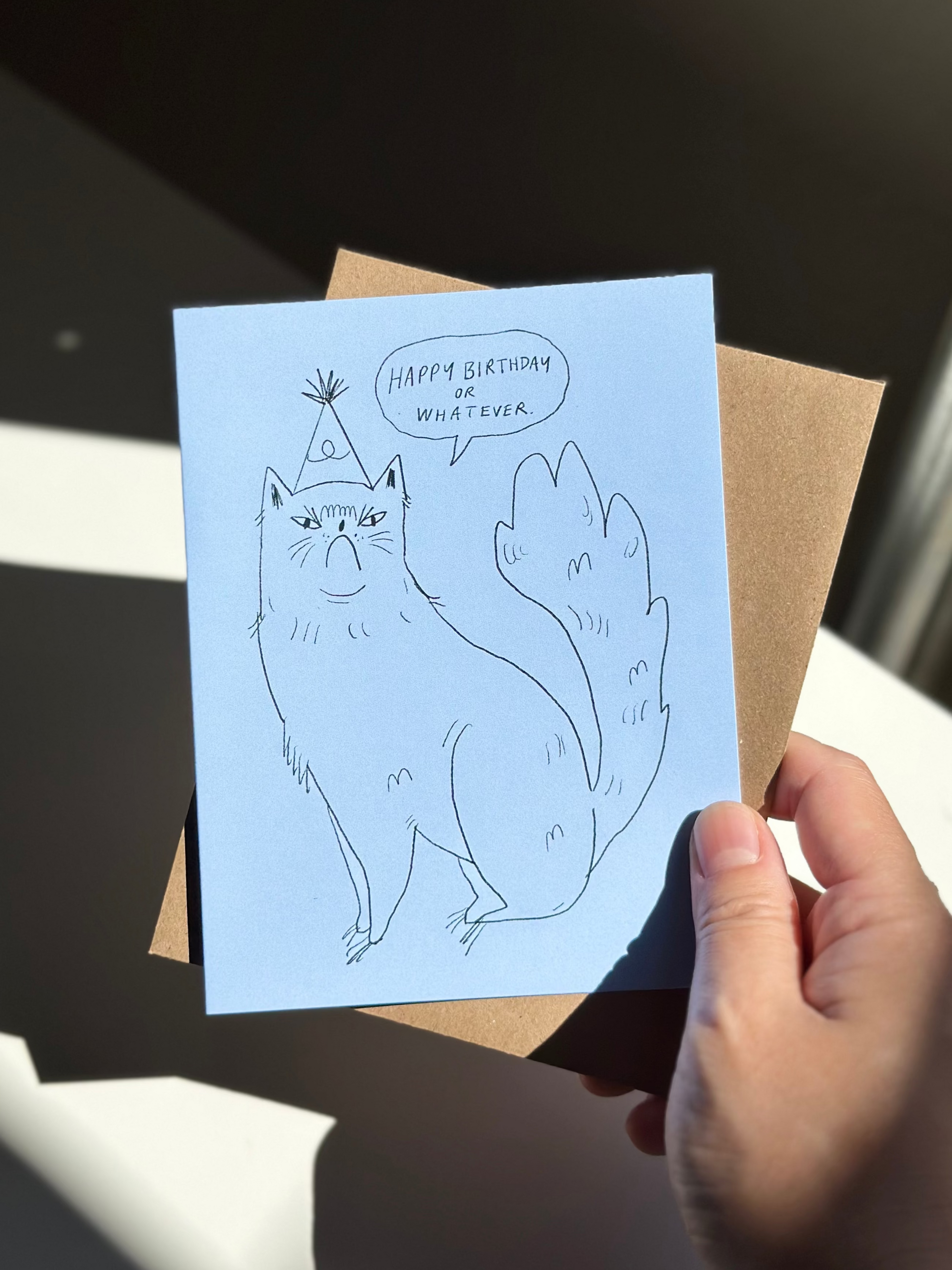 Funny cat birthday card with hand-drawn grumpy cat wearing party hat, saying ‘Happy Birthday or Whatever’ on blue card with brown kraft envelope.