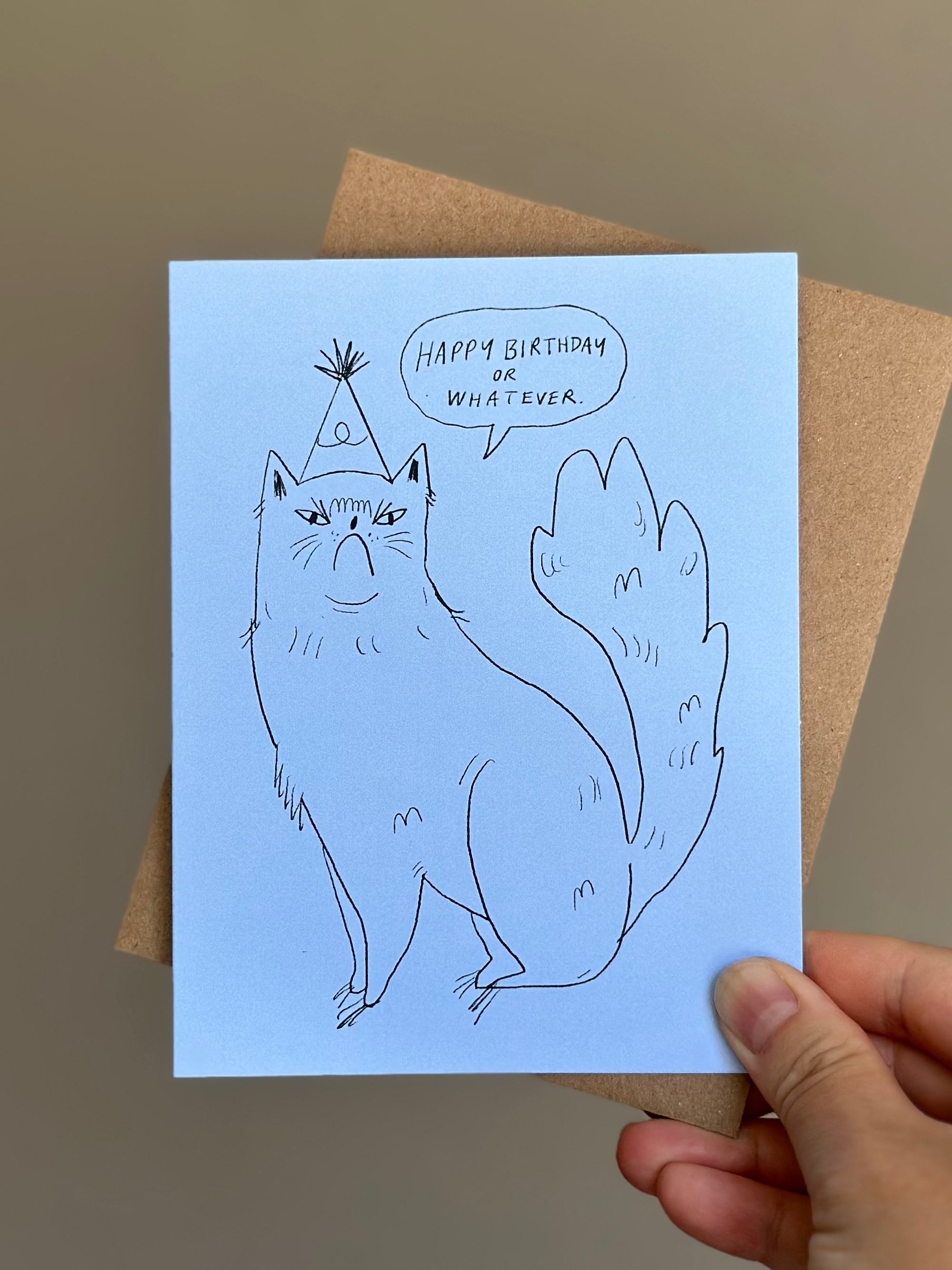 Funny cat birthday card with hand-drawn grumpy cat wearing party hat, saying ‘Happy Birthday or Whatever’ on blue card with brown kraft envelope.