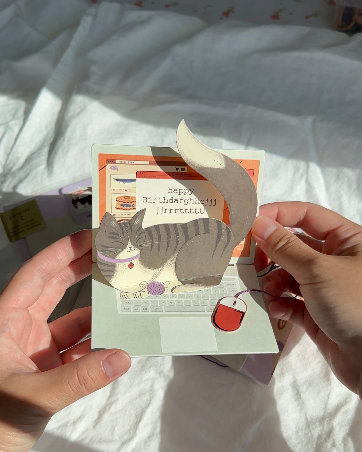 Person holding funny pop-up cat birthday card with 3D kitty typing on laptop keyboard.