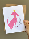 Funny greeting card with pink cartoon dog wearing a yellow crown and collar, captioned ‘You are one bad bitch,’ on white background with brown kraft envelope.